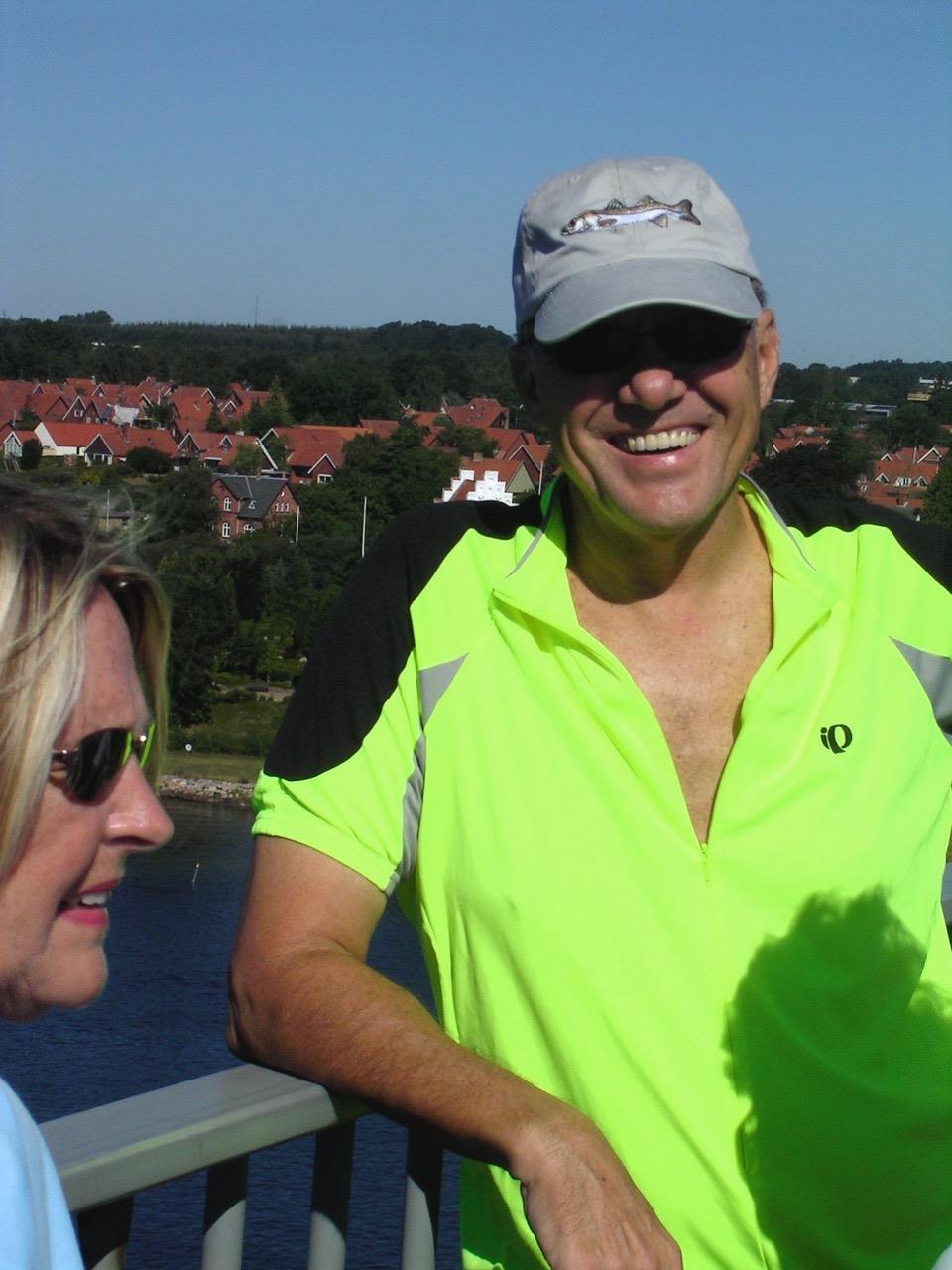 Two cyclists share a moment of joy near the water on a sunny day, dressed in bright gear.