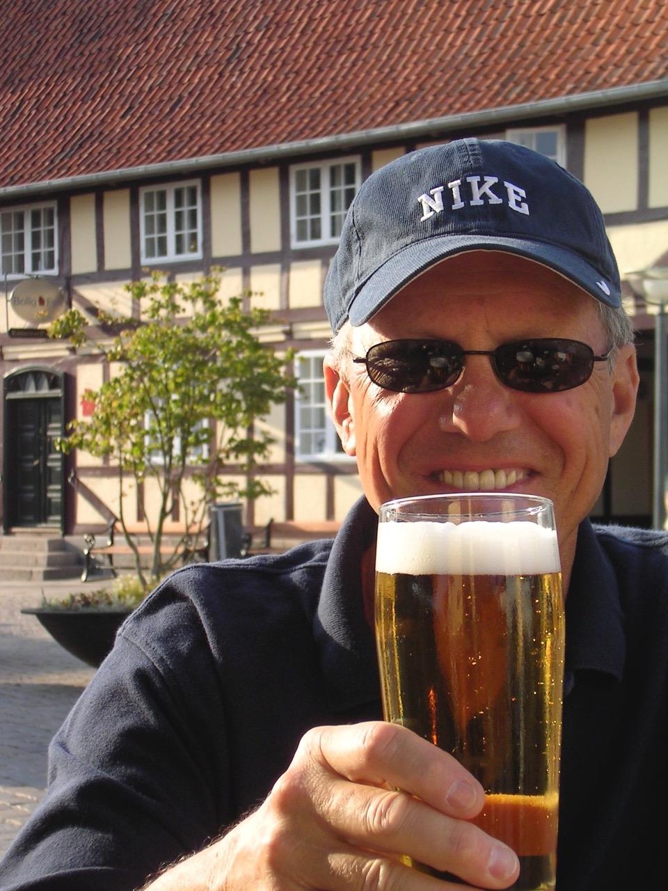 A man is smiling while holding a glass of beer in a beautiful outdoor space on a sunny day.
