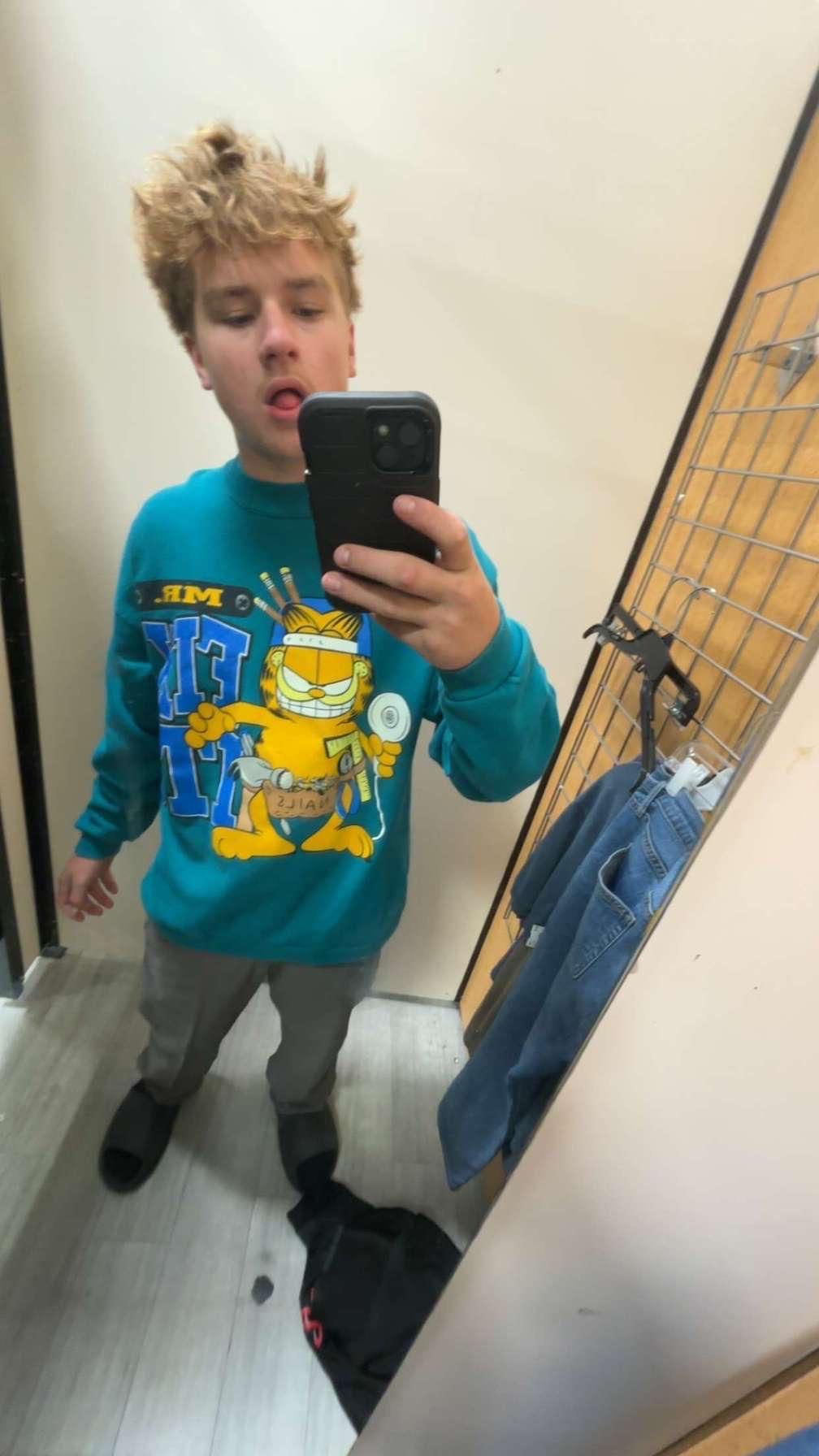 Young boy takes a selfie in a fitting room while wearing a colorful cartoon sweatshirt.