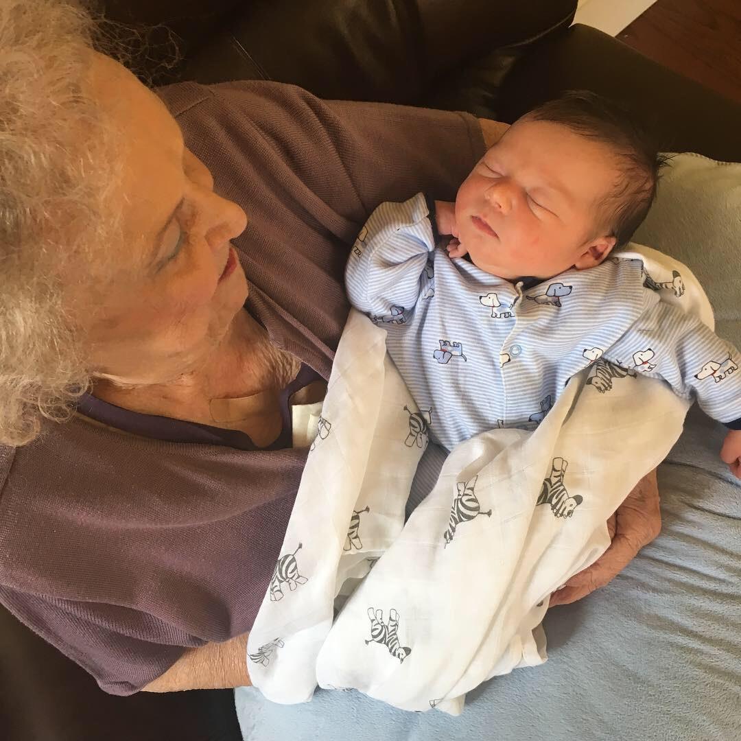 Elderly woman holds sleeping baby in soft blanket on a couch with warm colors in sunlight.