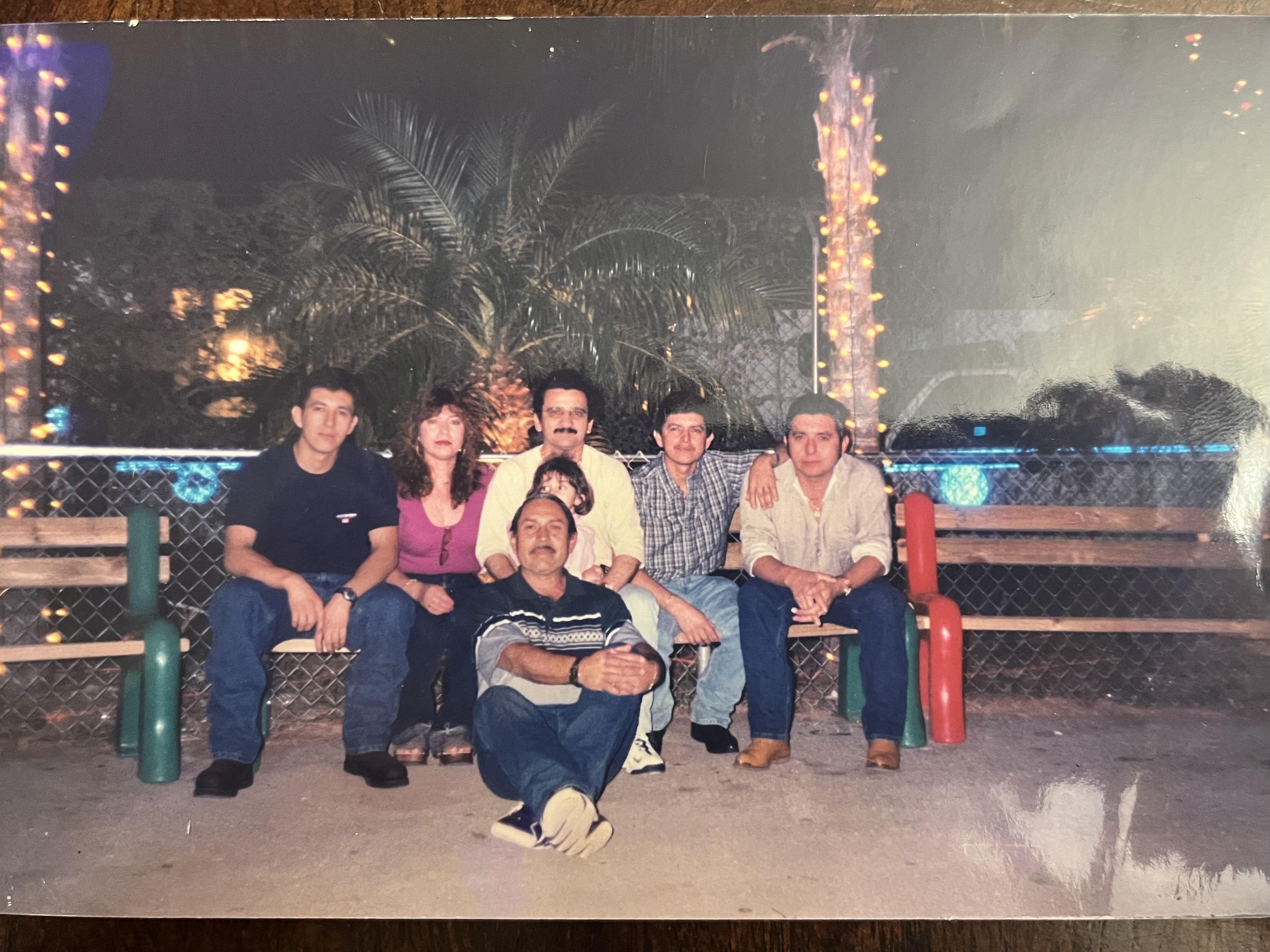 Friends gather on a bench under palm trees illuminated by festive lights in the evening.