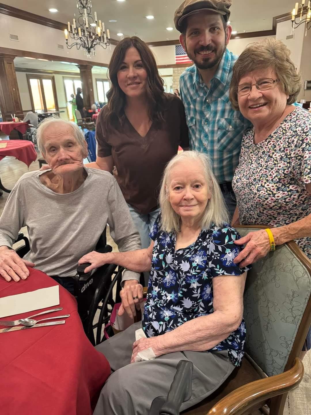 Elderly family members are seated together smiling at the dining table in a community center.