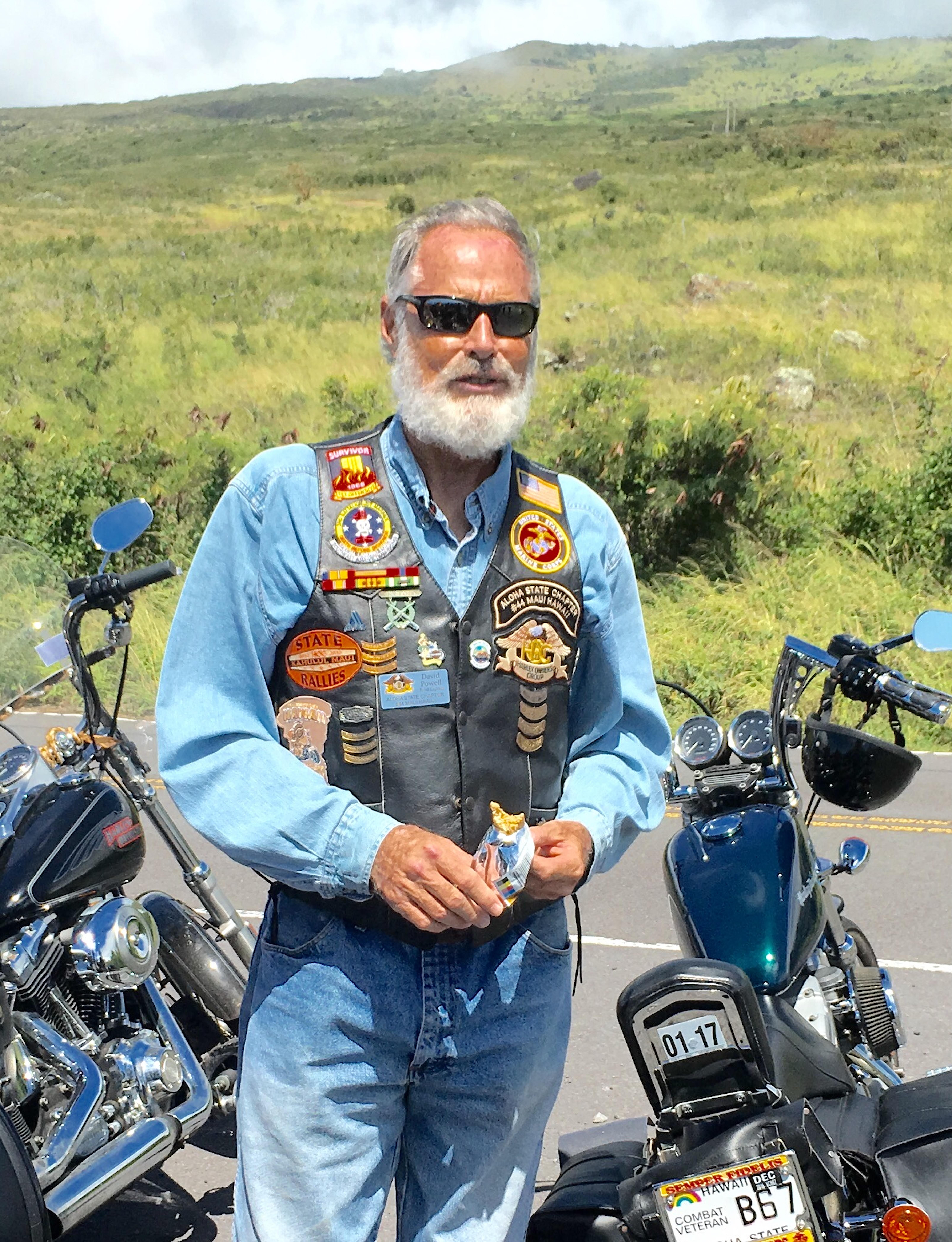 Biker with patches and sunglasses poses with motorcycles in a lush green landscape.