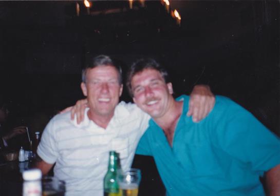 Two men sitting at a table with drinks and a bottle of beer