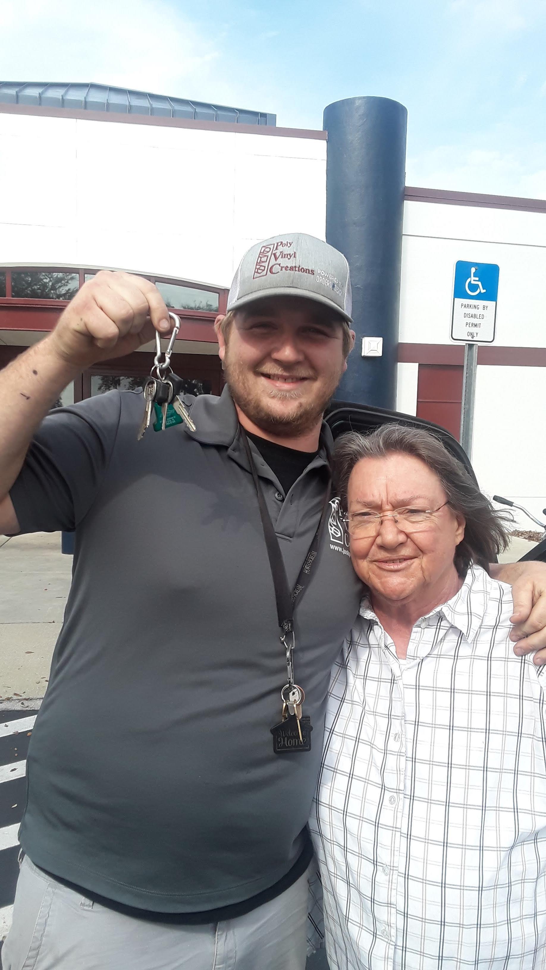 A man holds car keys high, celebrating his new car with a smiling older woman outside a dealership.