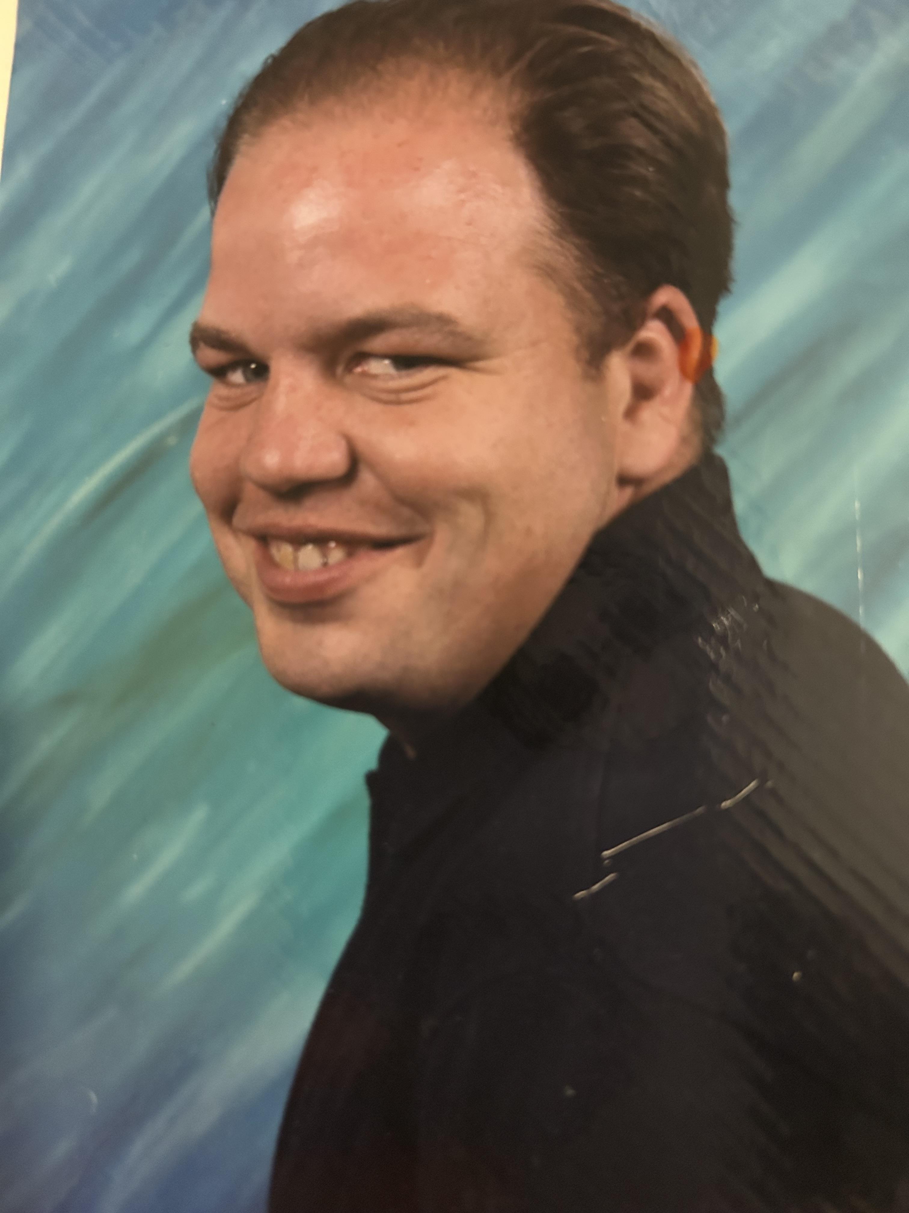 A man is smiling directly at the camera, dressed in a dark top.