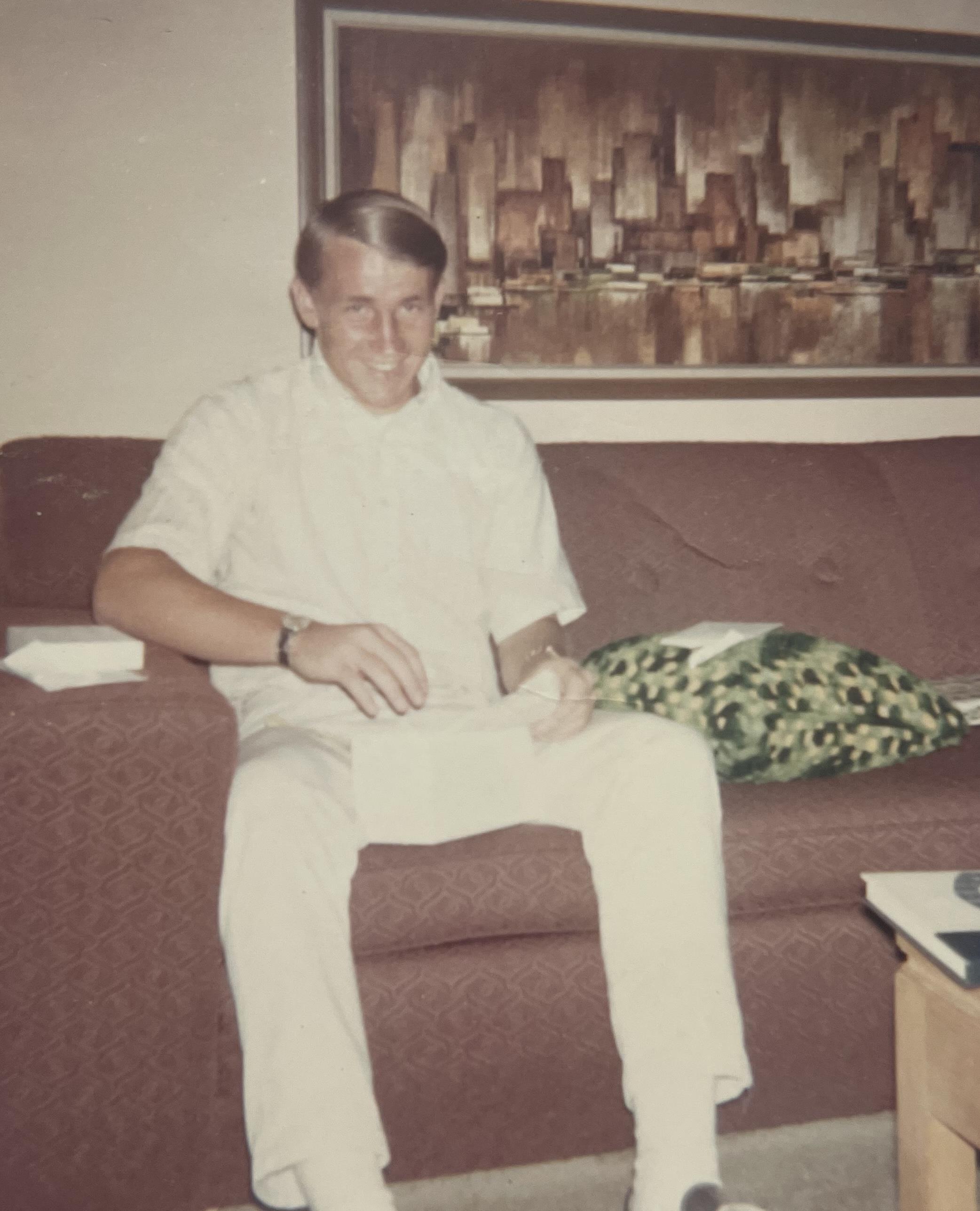 A man in white happily unwraps a package on a couch in a cozy living room.