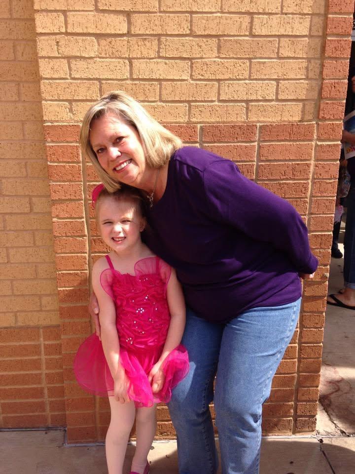 Girl in a pink dress smiles next to a woman by a brick building, sharing a joyful moment.