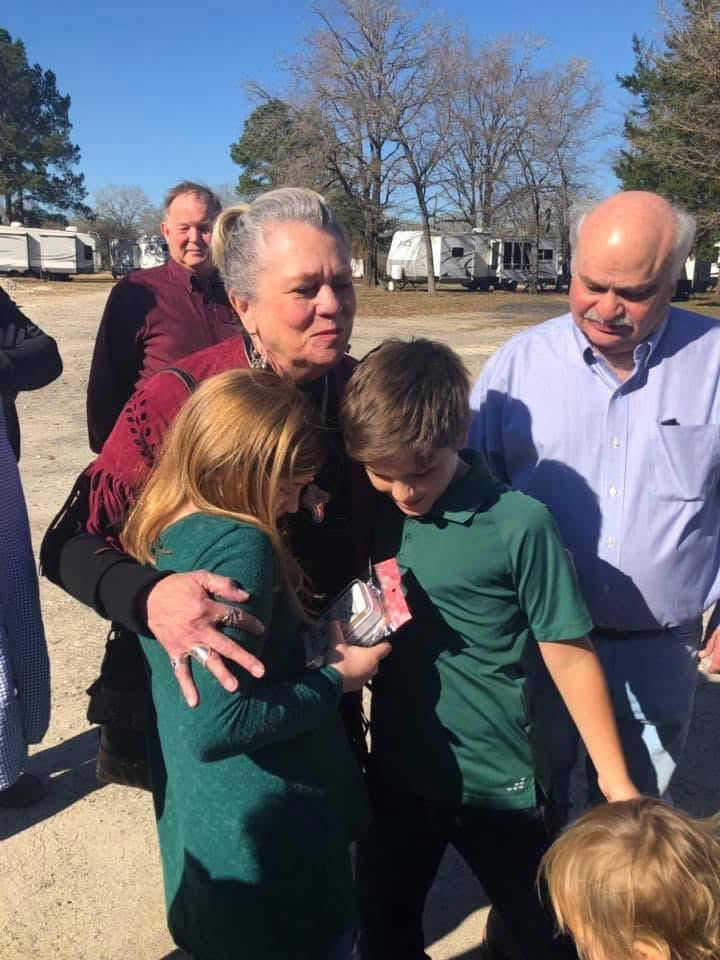 A woman joyously embraces two children while adults gather around in a sunny park.
