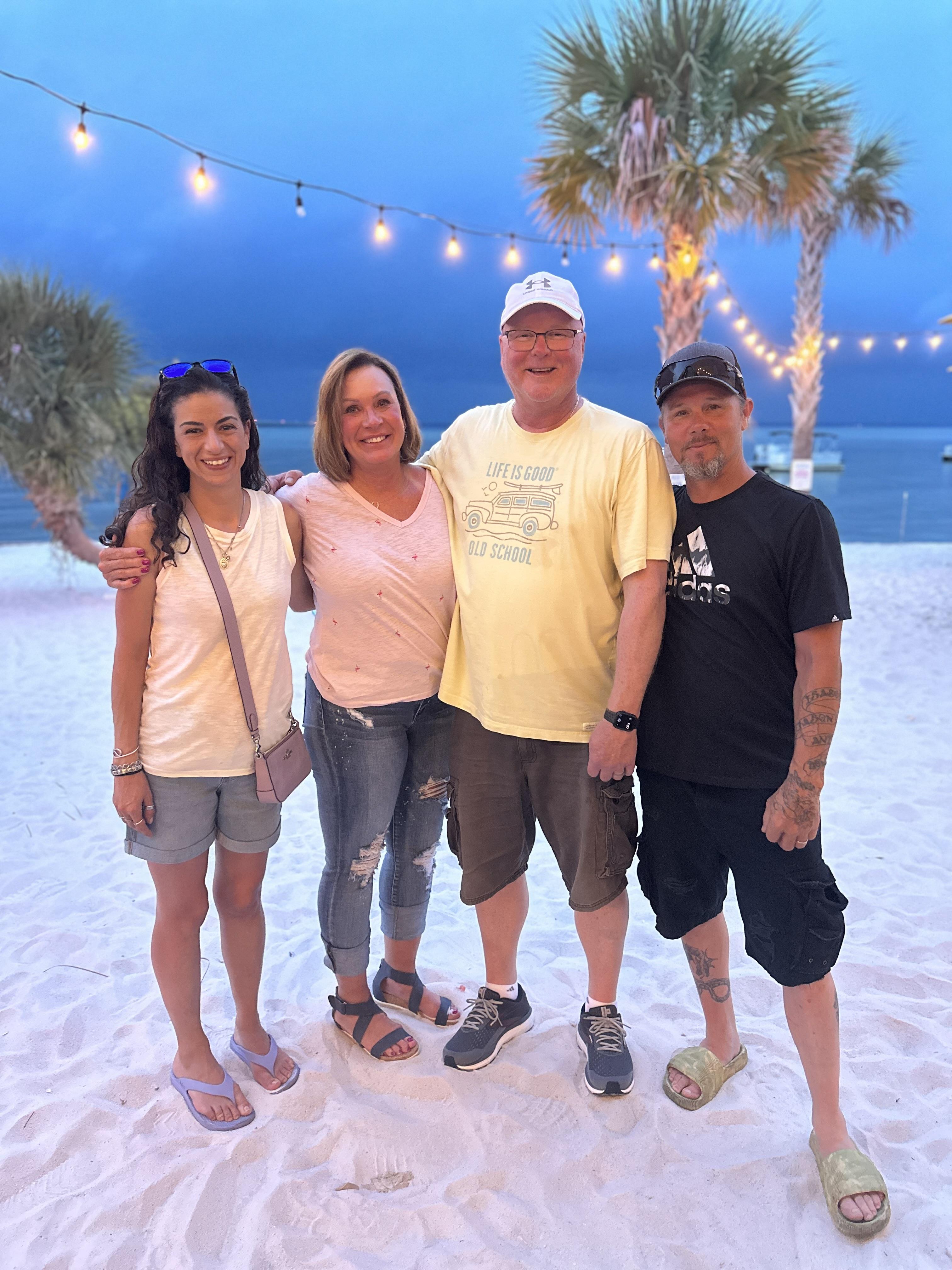 Friends stand together on a sandy beach surrounded by palm trees and evening lights.