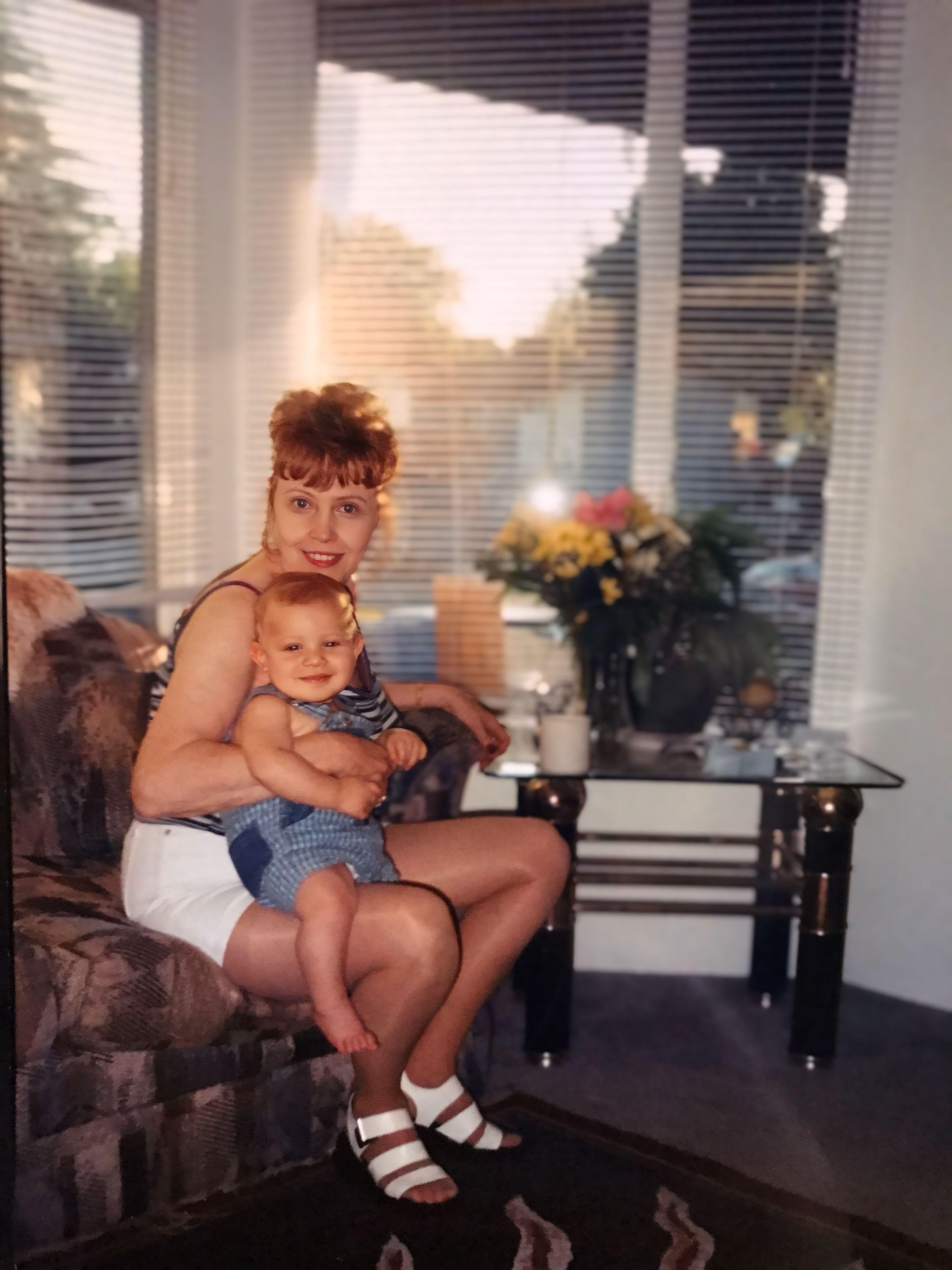 A joyful mother sits on a couch with her smiling baby during a warm evening, surrounded by sunlight.