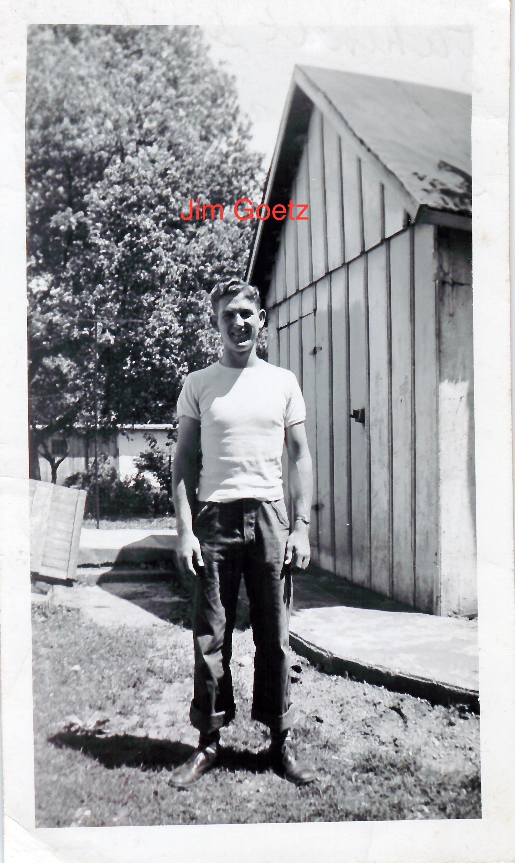 A young man stands confidently in a yard beside a wooden shed, wearing a simple t-shirt and pants.