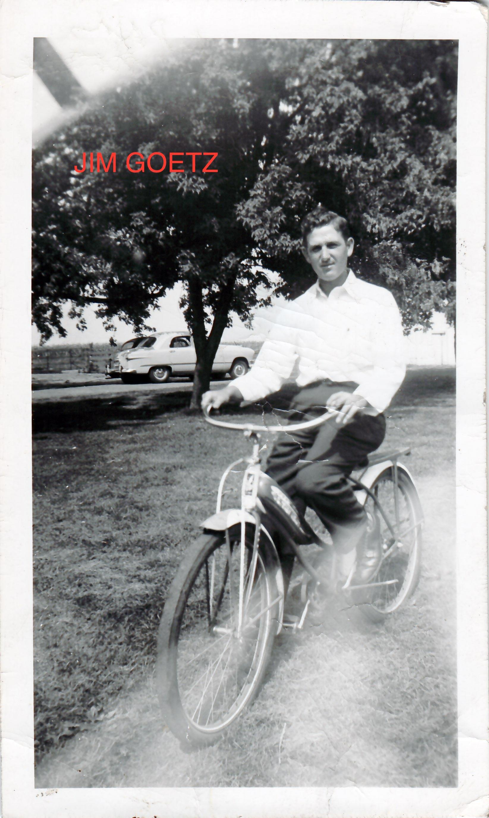 A man enjoys a sunny day riding a bicycle in a park, surrounded by trees and vintage cars.