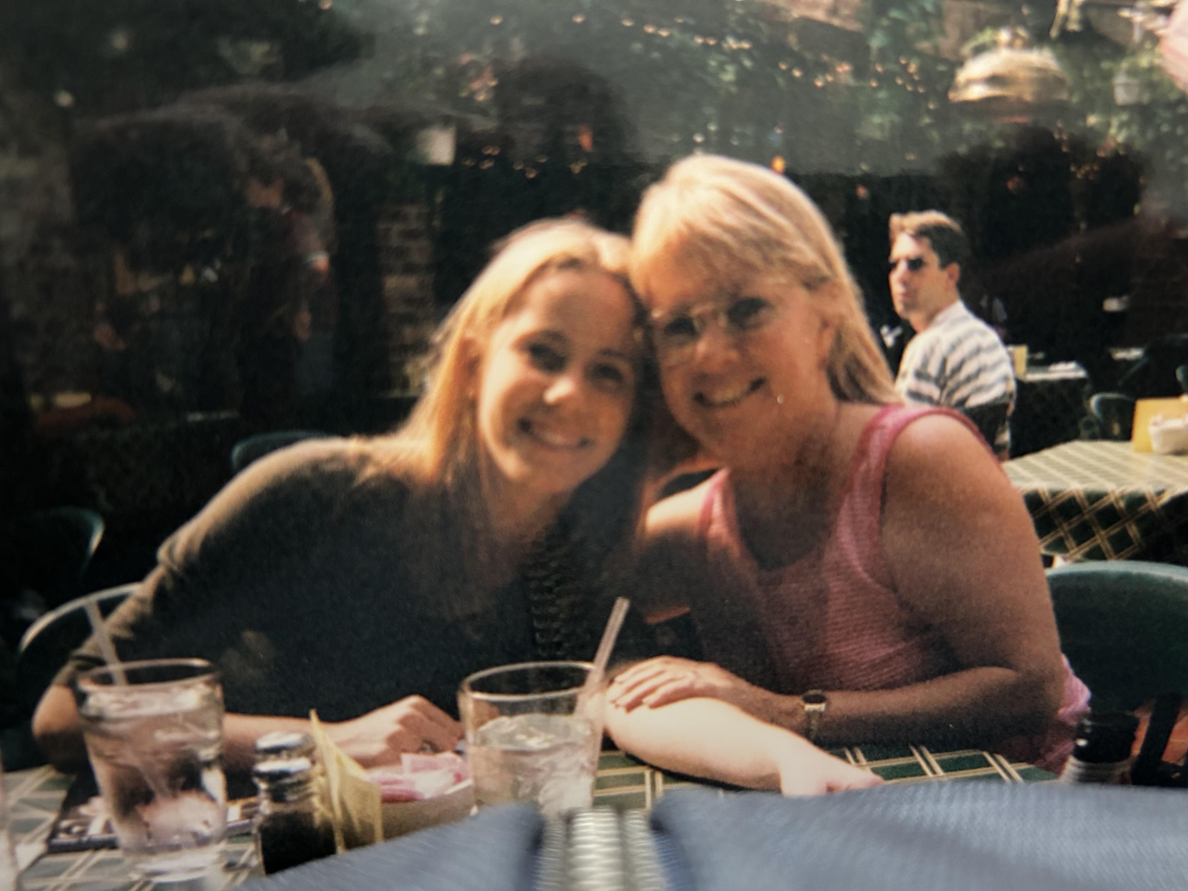 Two friends smile warmly together while sitting at a table in a bustling restaurant.