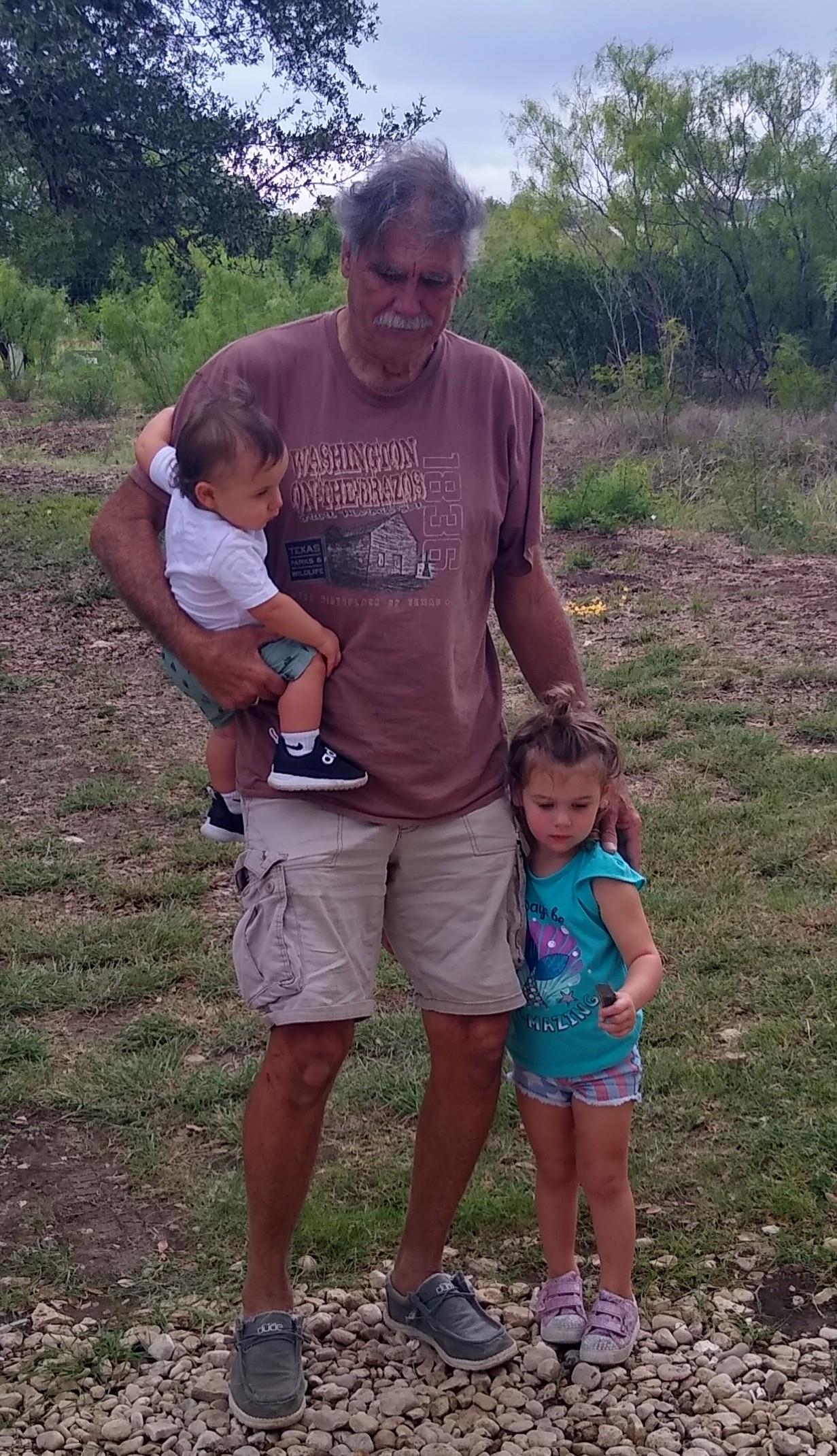 An elderly man stands on grassy ground holding one child and watching another play nearby.