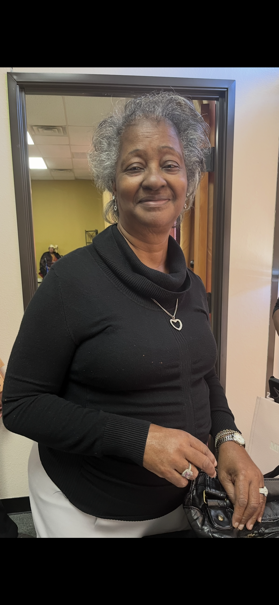A woman with gray hair and a welcoming smile stands in a community center, enjoying the gathering.