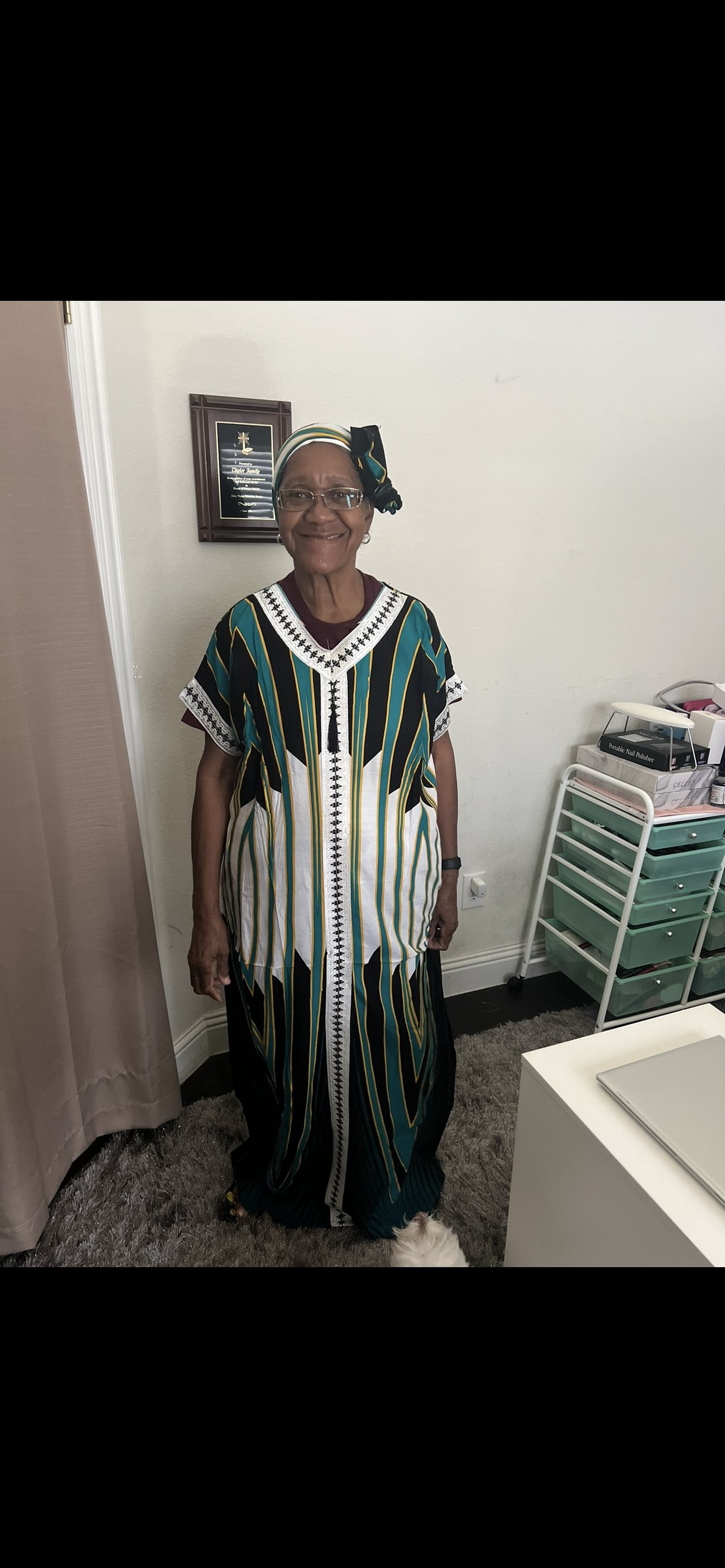 An elderly woman in traditional attire smiles warmly in her decorated home.