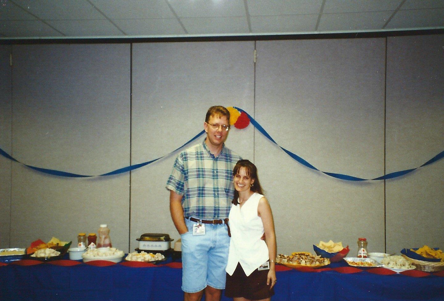 Two people stand together smiling in front of a festive food table with colorful decorations.