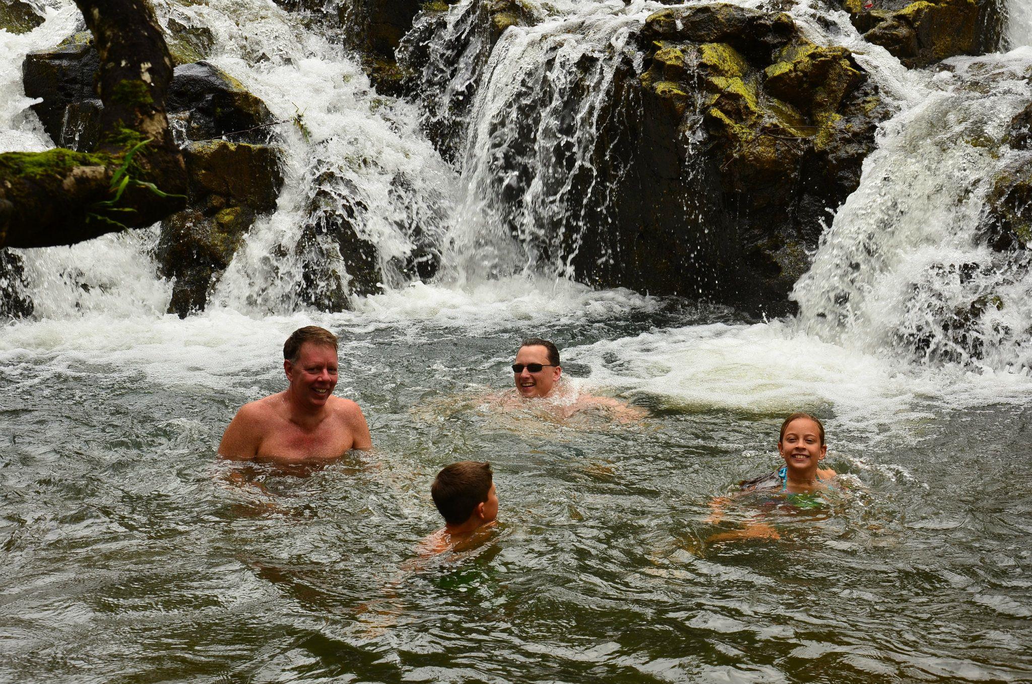 A family swims joyfully in a refreshing waterfall pool surrounded by lush greenery.