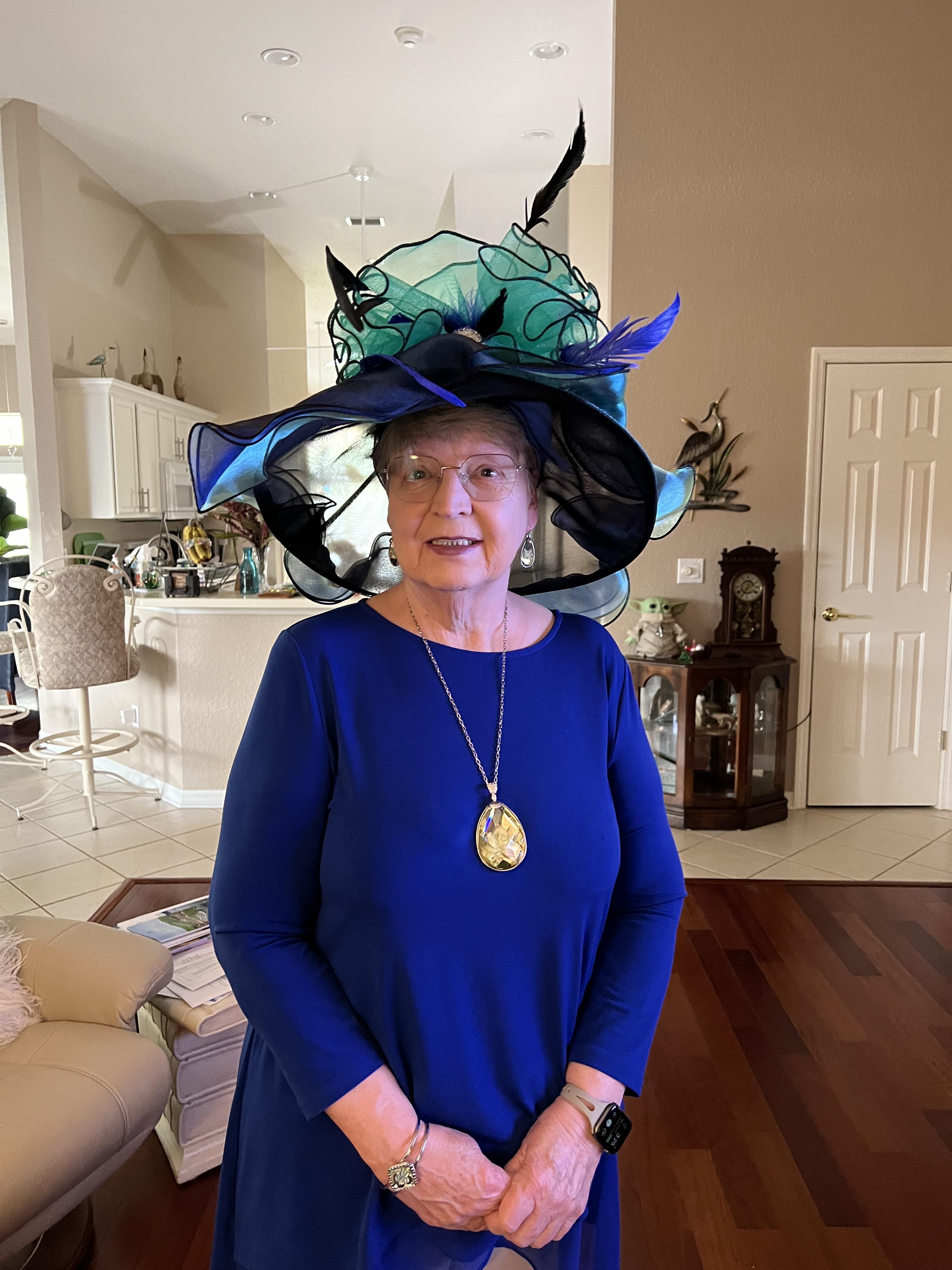 A well-dressed older woman poses proudly in a vibrant living room, showcasing her unique hat.