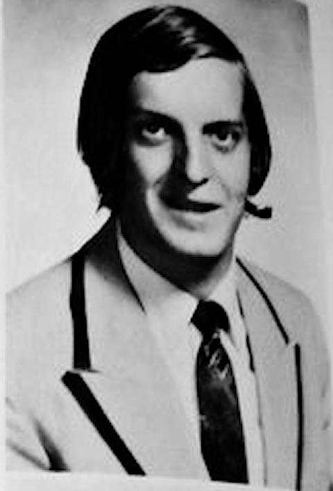 A man smiles while wearing a headset, dressed in a suit and tie, posing for a formal portrait.