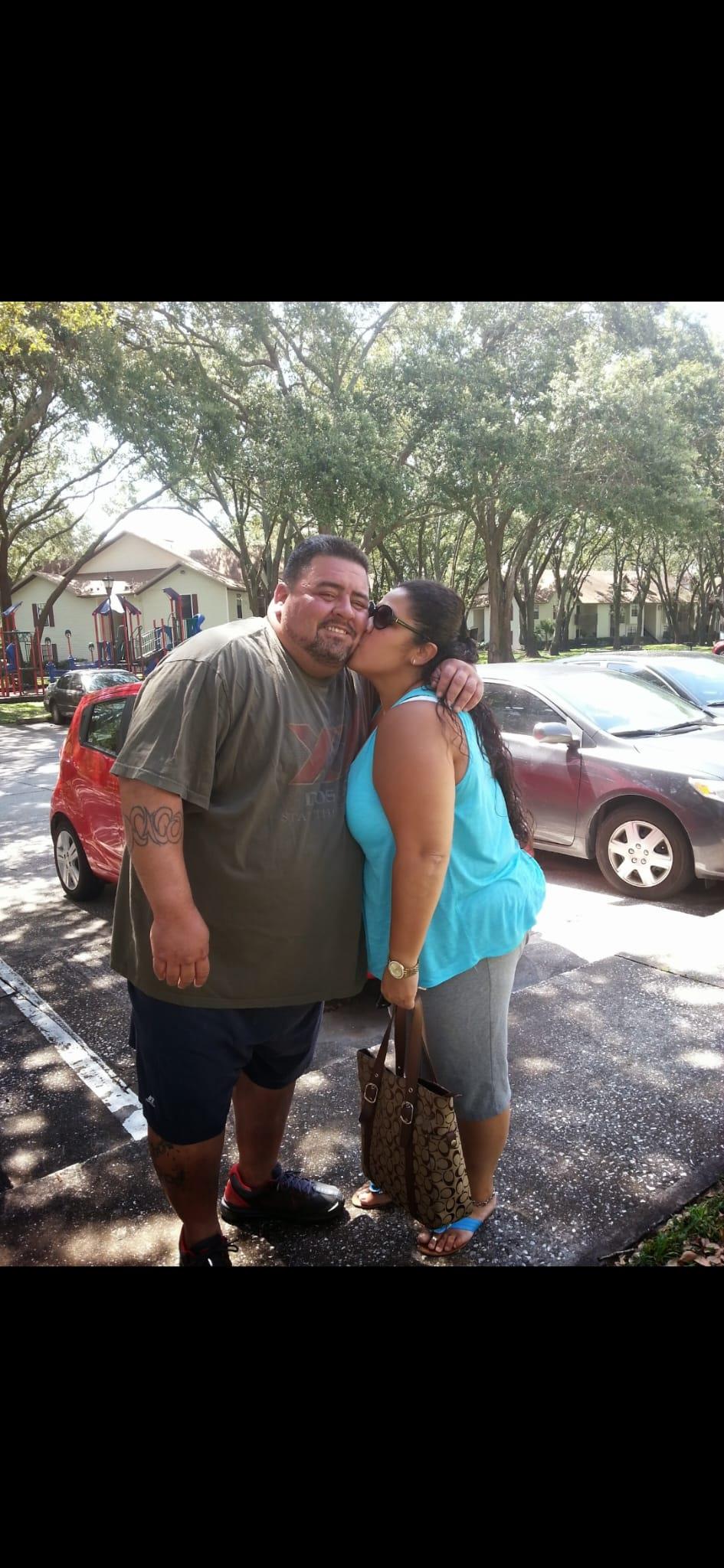 Two people smile and embrace in a parking lot filled with cars on a bright day.