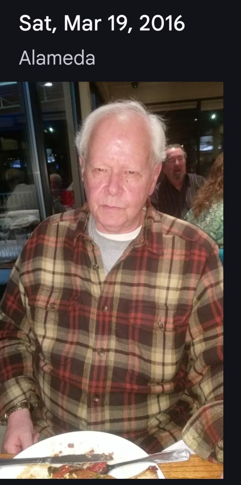 Mature man with silver hair wearing a plaid shirt sits at a table, looking thoughtful.