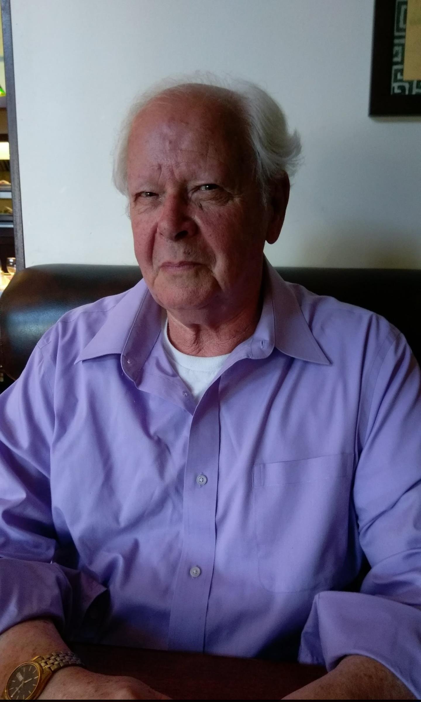 An older man with gray hair sits at a table with a serious expression, wearing a purple shirt.
