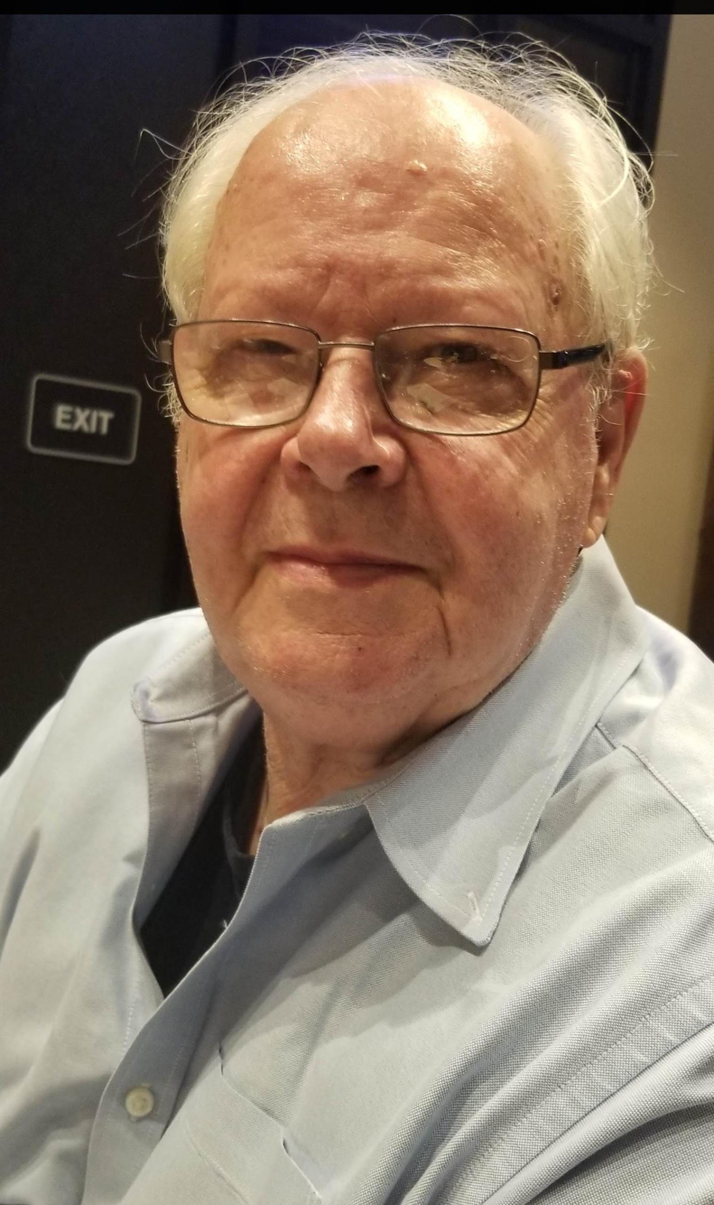 An elderly man wearing glasses sits calmly with a serious expression in a cozy indoor environment.