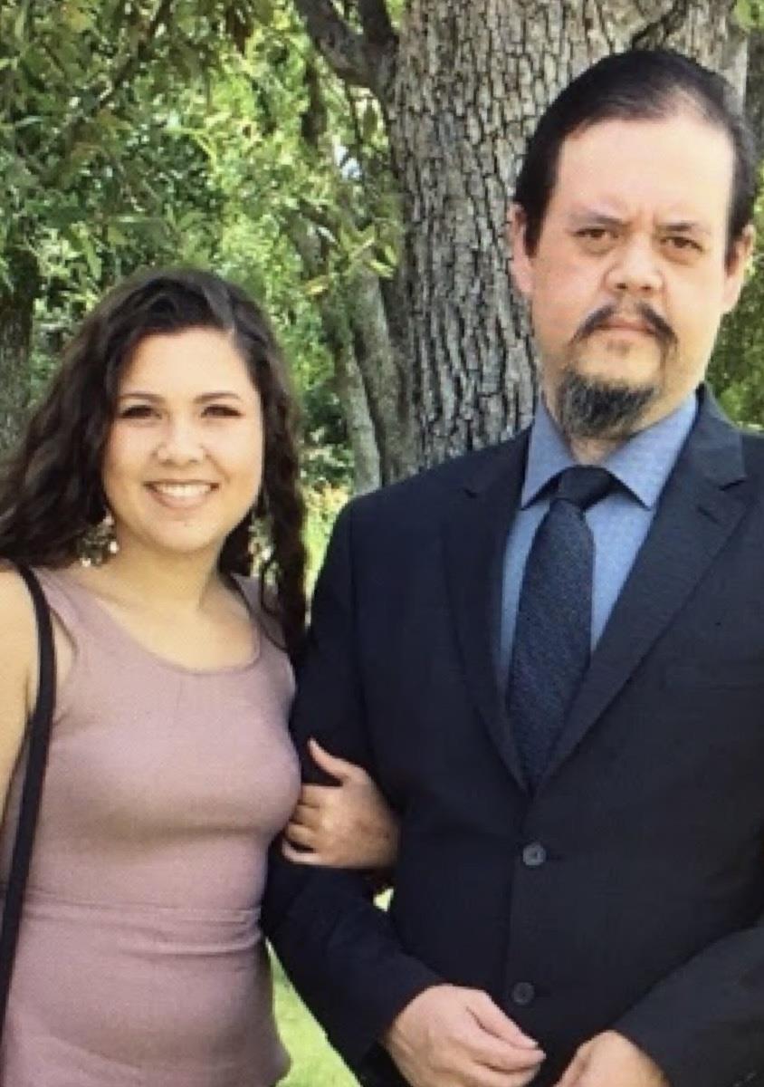 Two individuals are smiling and standing close to each other in a green outdoor setting.