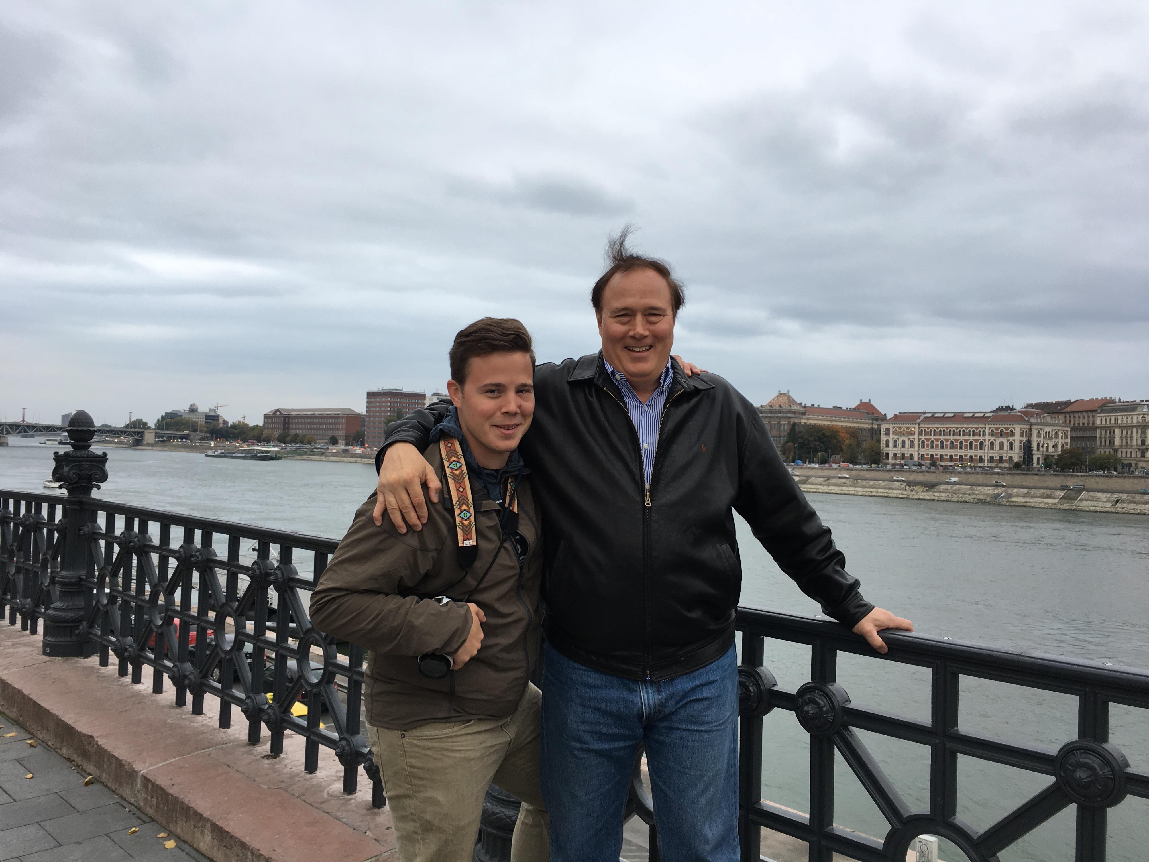Two friends stand by the river in Budapest, smiling against a backdrop of cloudy skies.