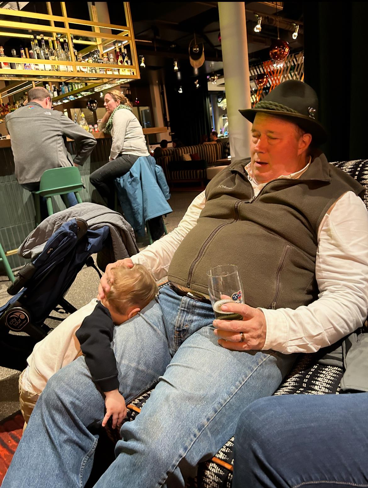 Man relaxes in bar while child curiously explores his surroundings, capturing a heartwarming moment.