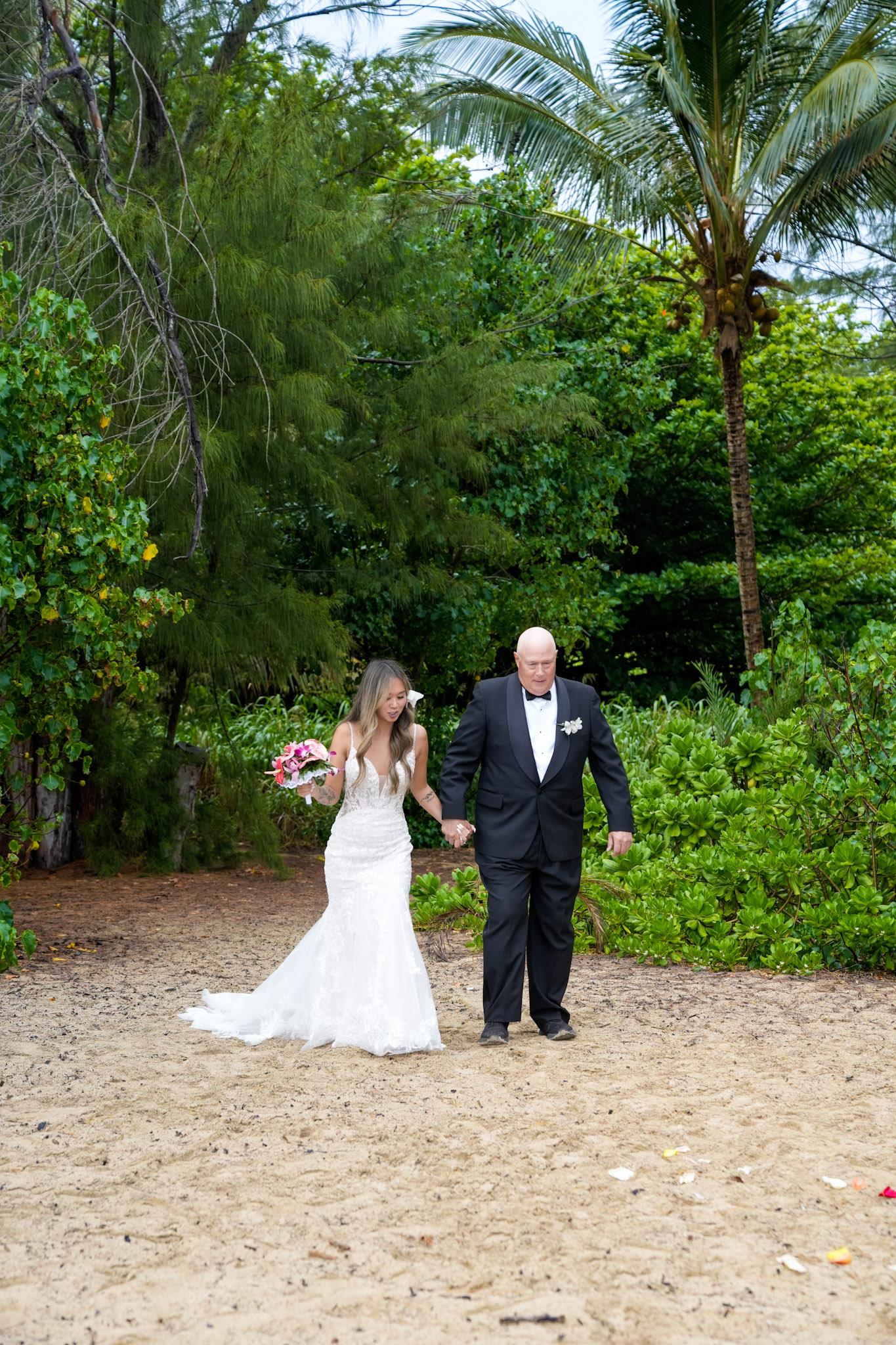 Bride in white gown and groom in black suit stroll along sandy beach, celebrating their special day.