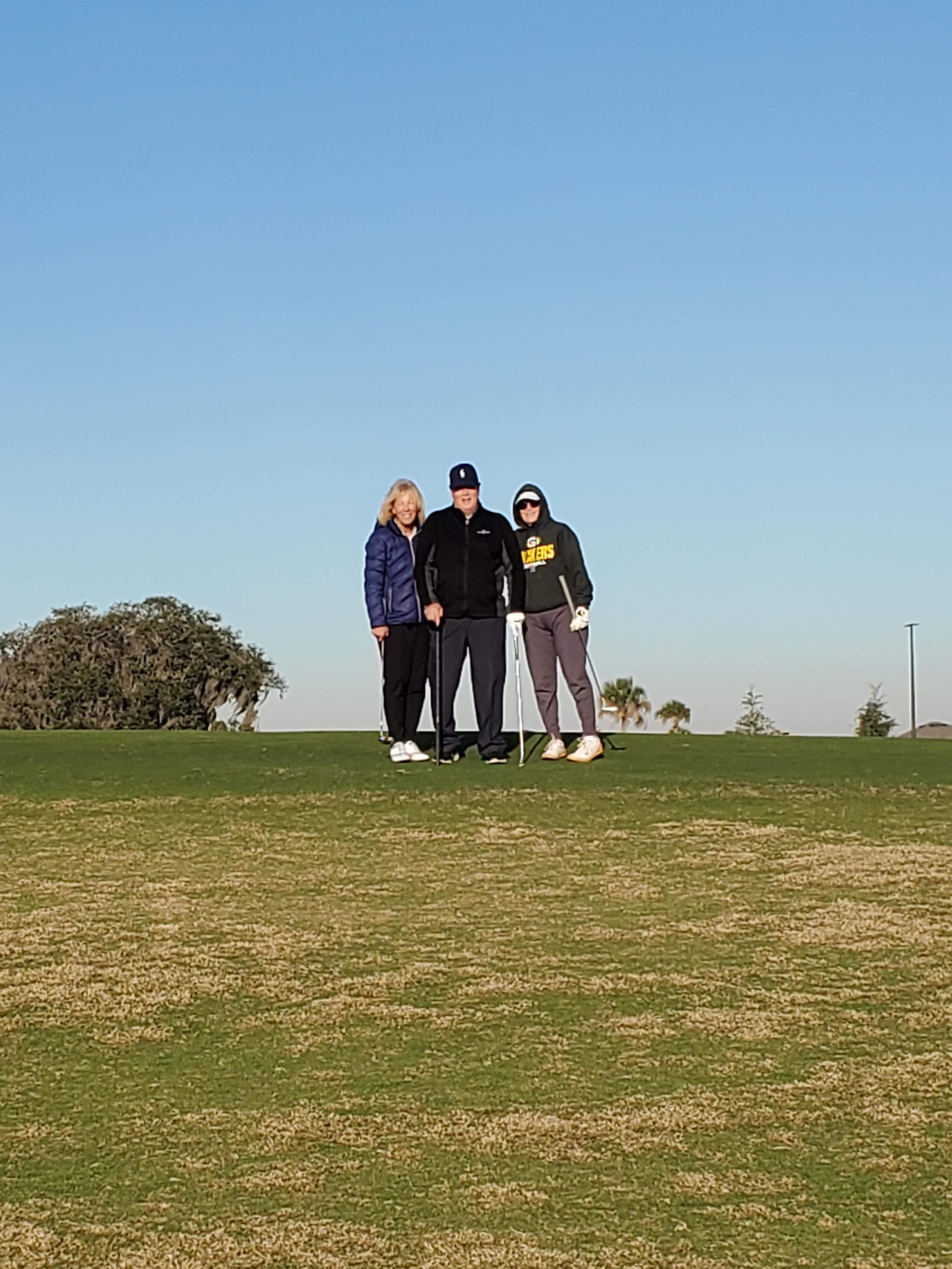 Three individuals stand on a golf course, enjoying a sunny day while holding golf clubs.