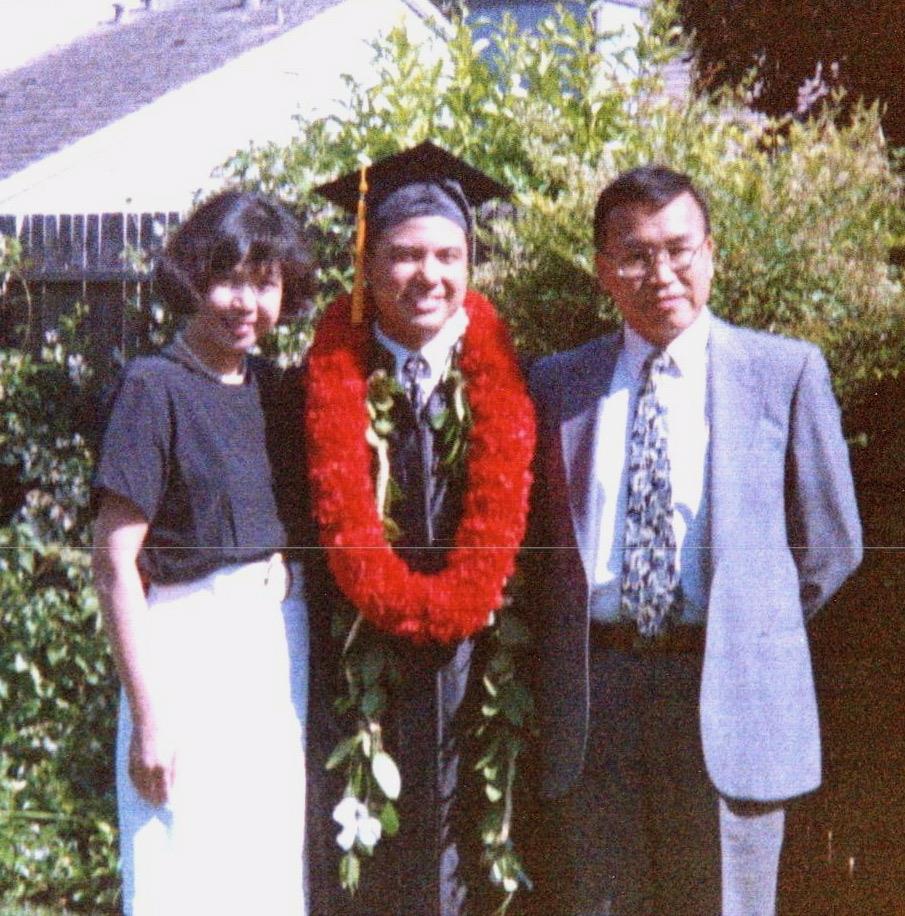 A proud graduate stands between his parents wearing a graduation cap and lei, smiling warmly.