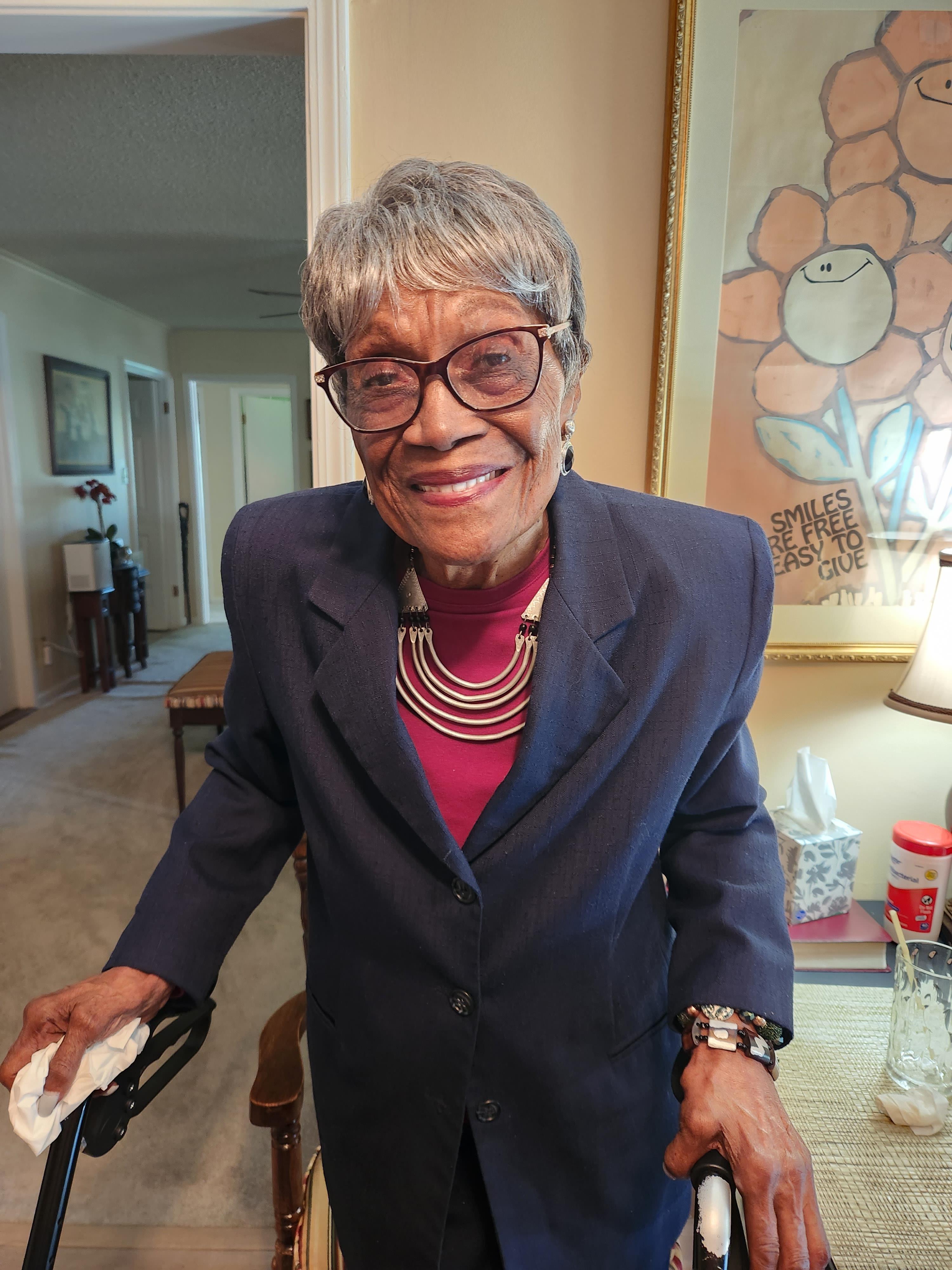 An elderly woman in a blue blazer stands in a warm living space, smiling and looking cheerful.