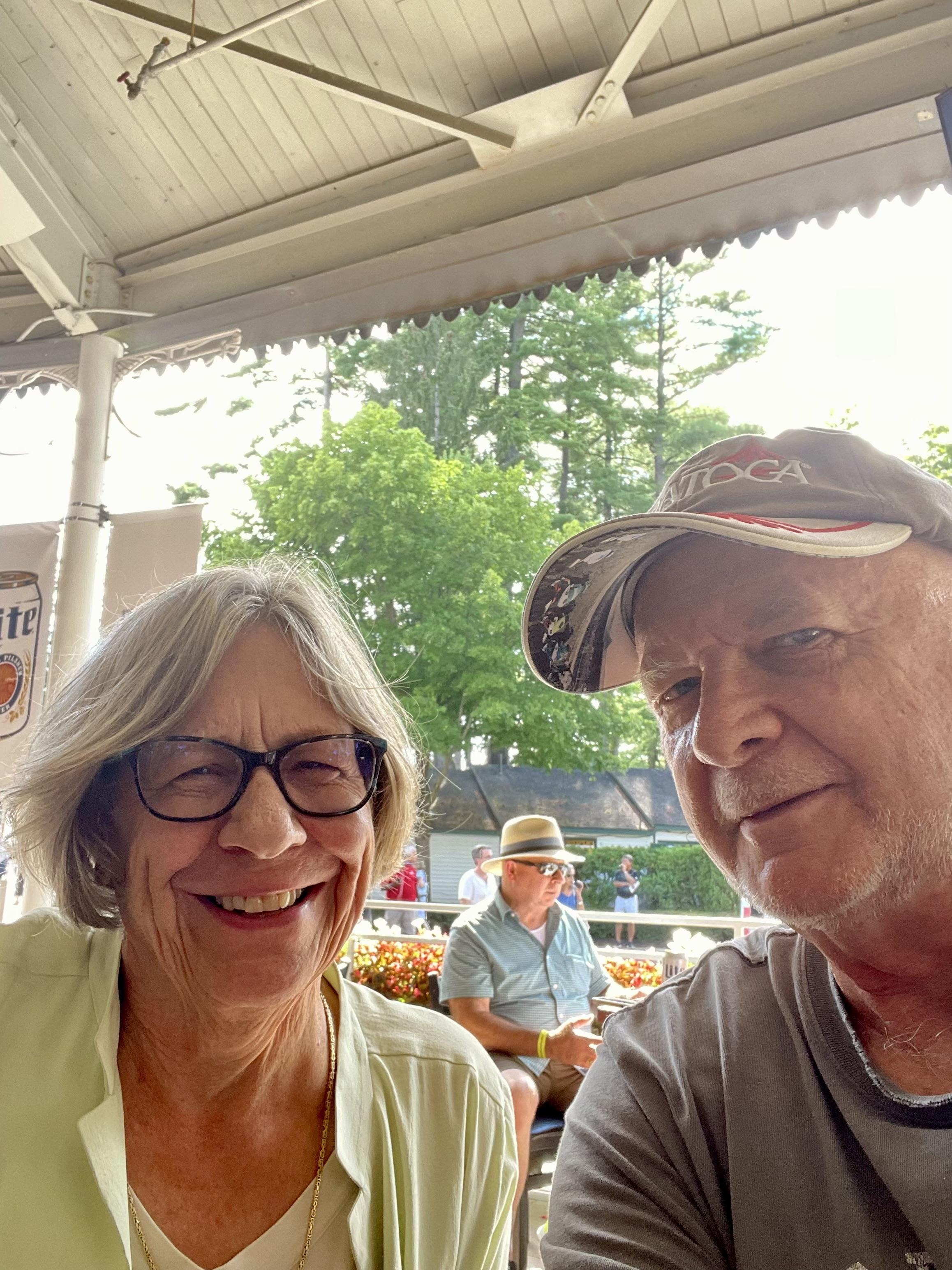 Two seniors smile happily together at an outdoor café while others relax in the background.