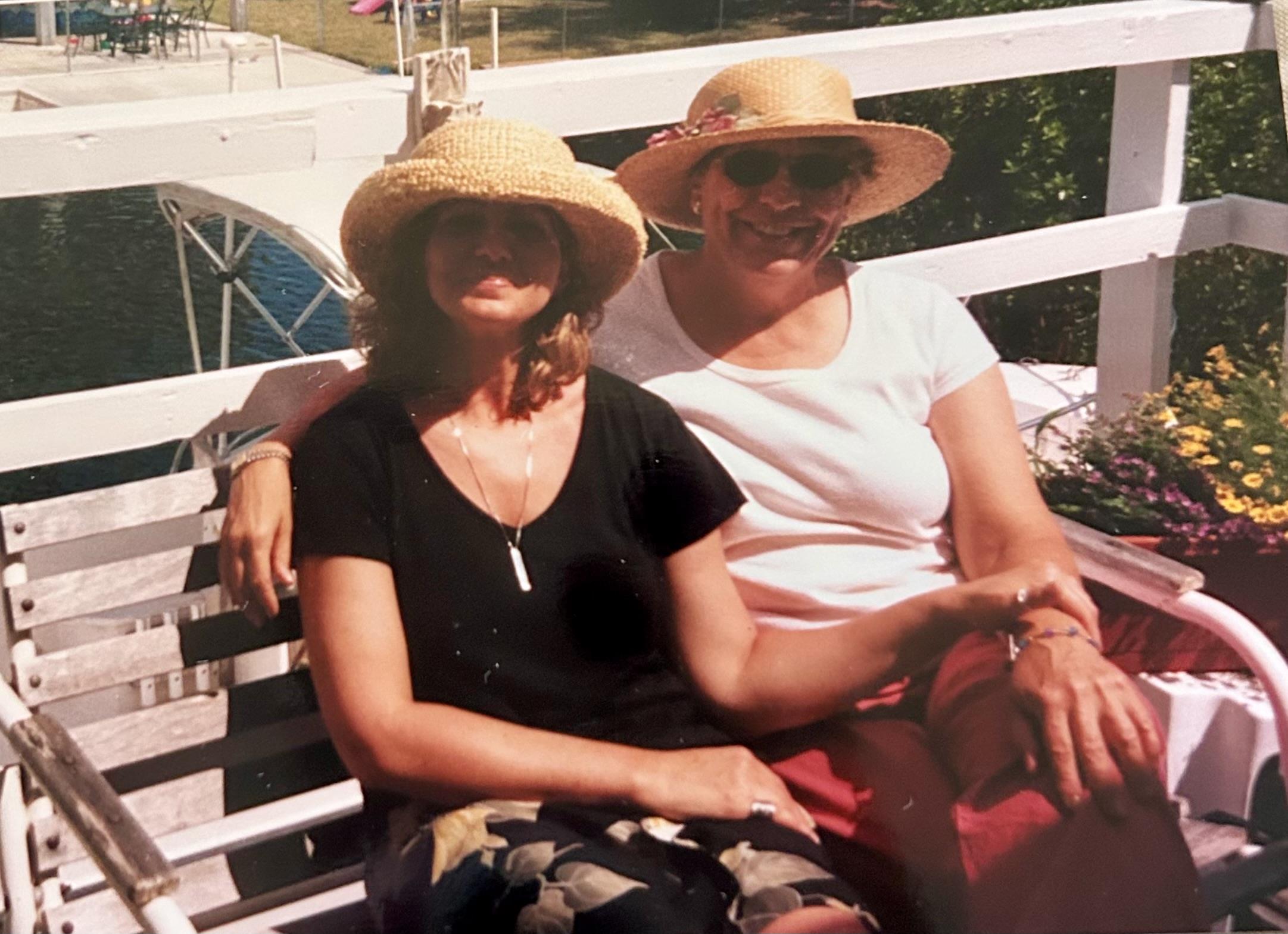 Two women sit on a bench in summer attire, smiling under the sun by the lake.