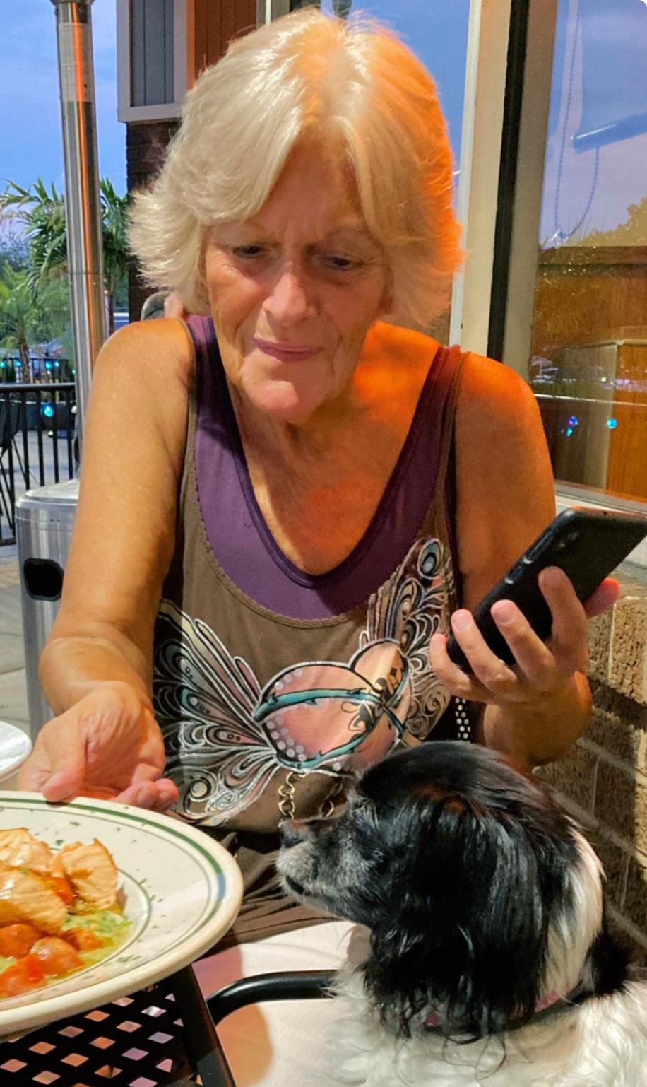 A woman sits at a restaurant table, focused on her phone while sharing food with her dog.