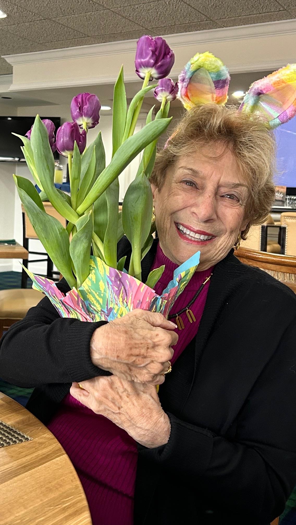 Older woman smiles widely while holding a vibrant bouquet of tulips in a decorated pot.