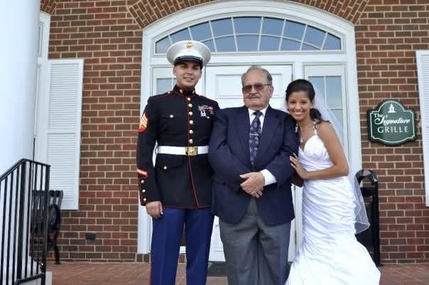 A Marine proudly poses with his grandfather and bride outside a venue, celebrating.