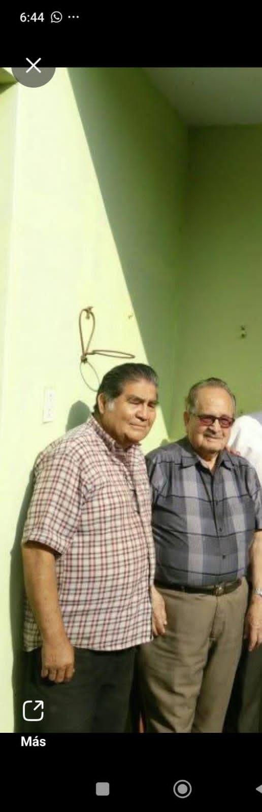 Two men stand closely together, smiling at the camera while enjoying a sunny day outdoors.