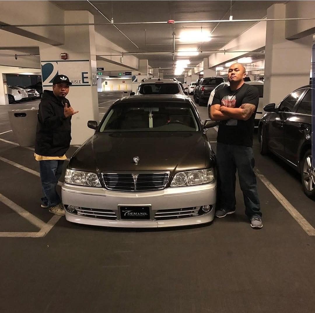 Two men show attitude while standing next to a modified car in a dim underground parking lot.