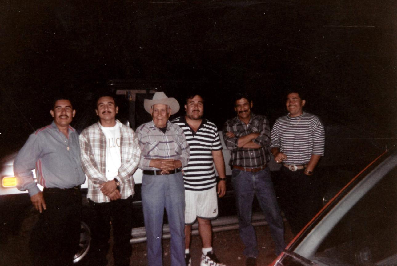 Six men stand together, smiling under dim light during a nighttime gathering in a remote area.