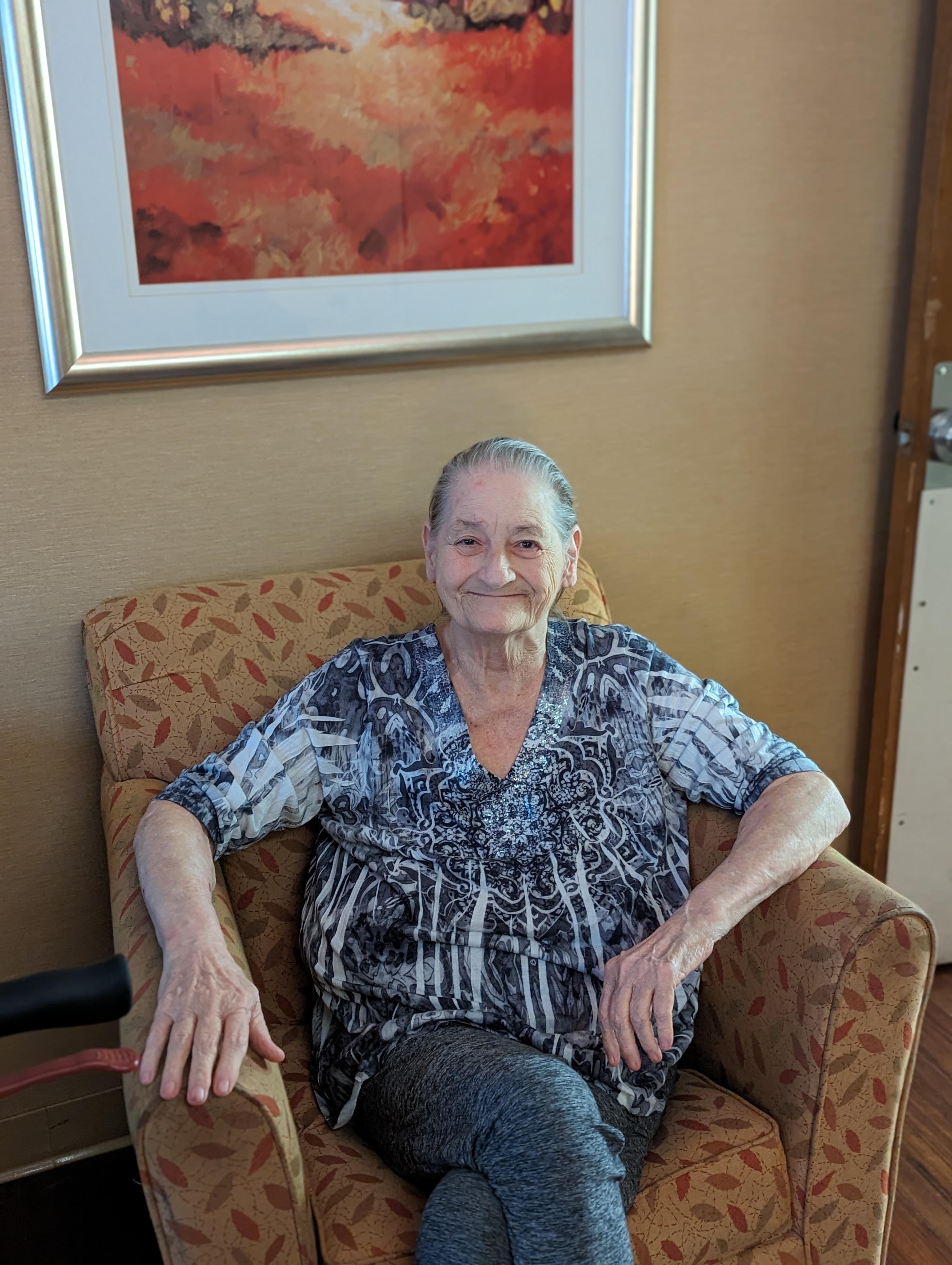 Older woman sits in a patterned chair, looking happy and relaxed in a bright indoor space.