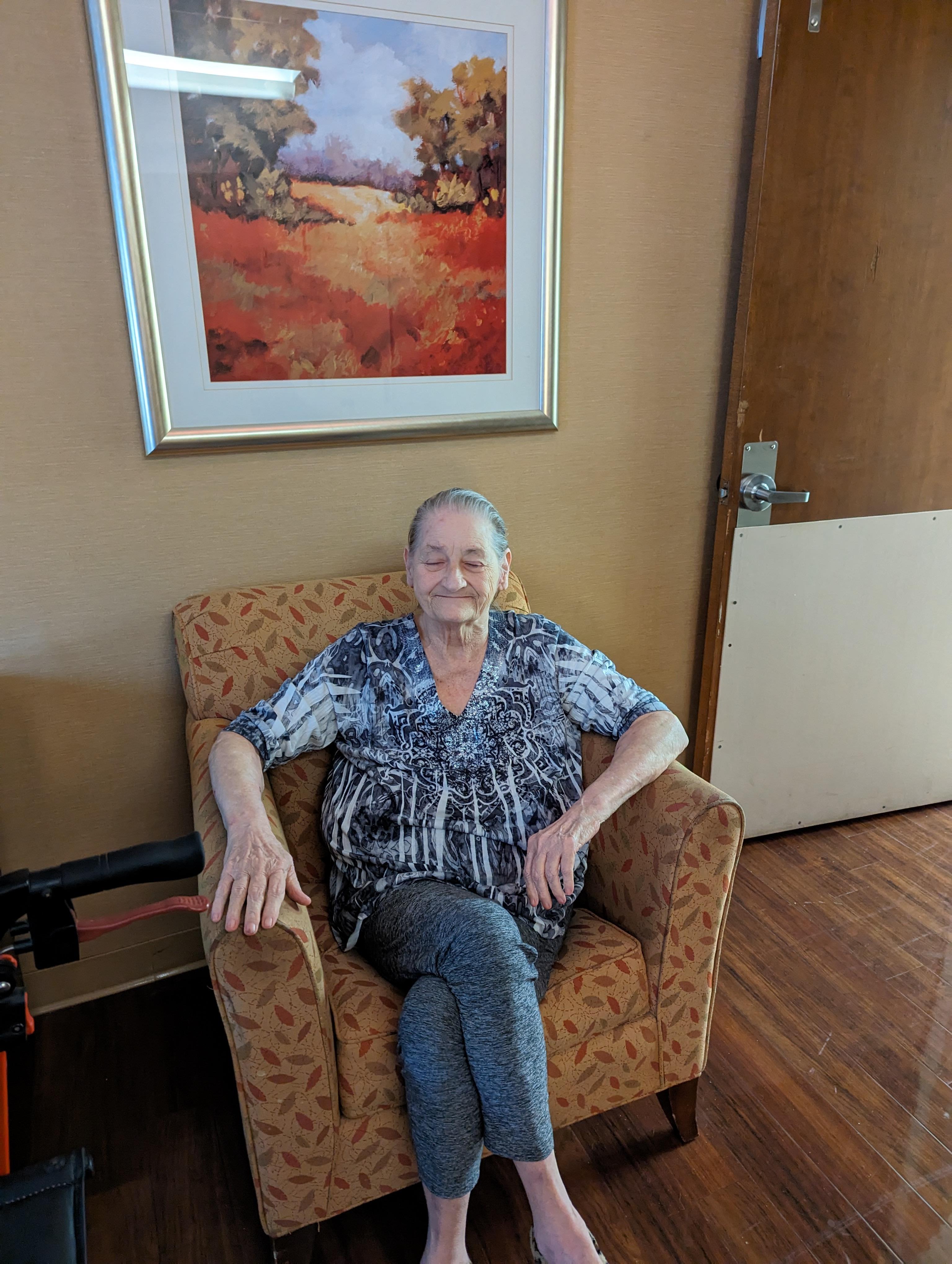 A senior woman sits comfortably in a chair, smiling softly in a welcoming room with decor.