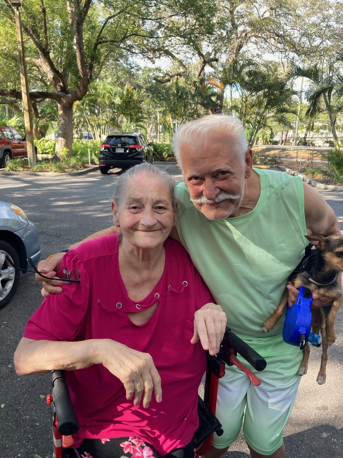 Seniors share a joyful moment outside, accompanied by their small dog, amidst lush greenery.