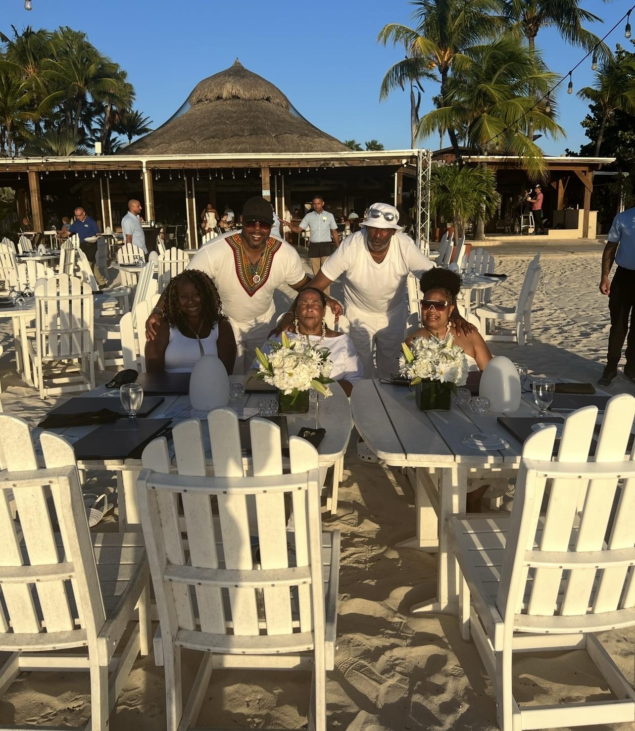 A group of friends in white attire gathers on a sandy beach, enjoying a lively outdoor celebration.
