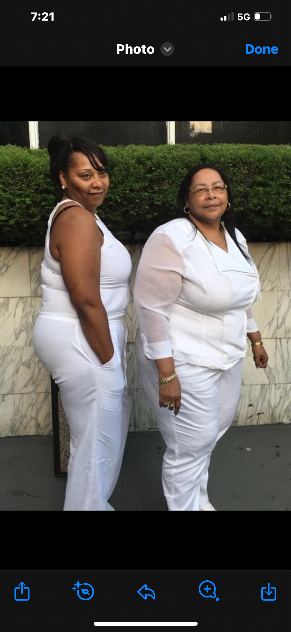 Two women smile while dressed in white outfits, standing close together outside against greenery.