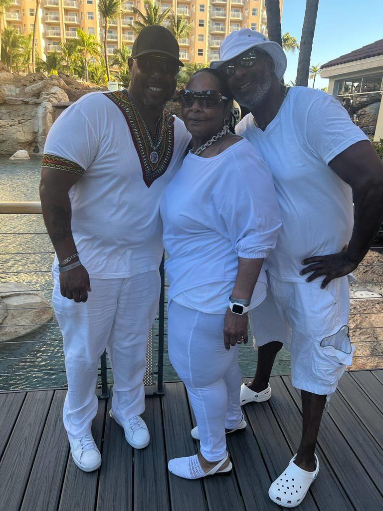 Three friends dressed in all white stand together, smiling at a resort by the water.